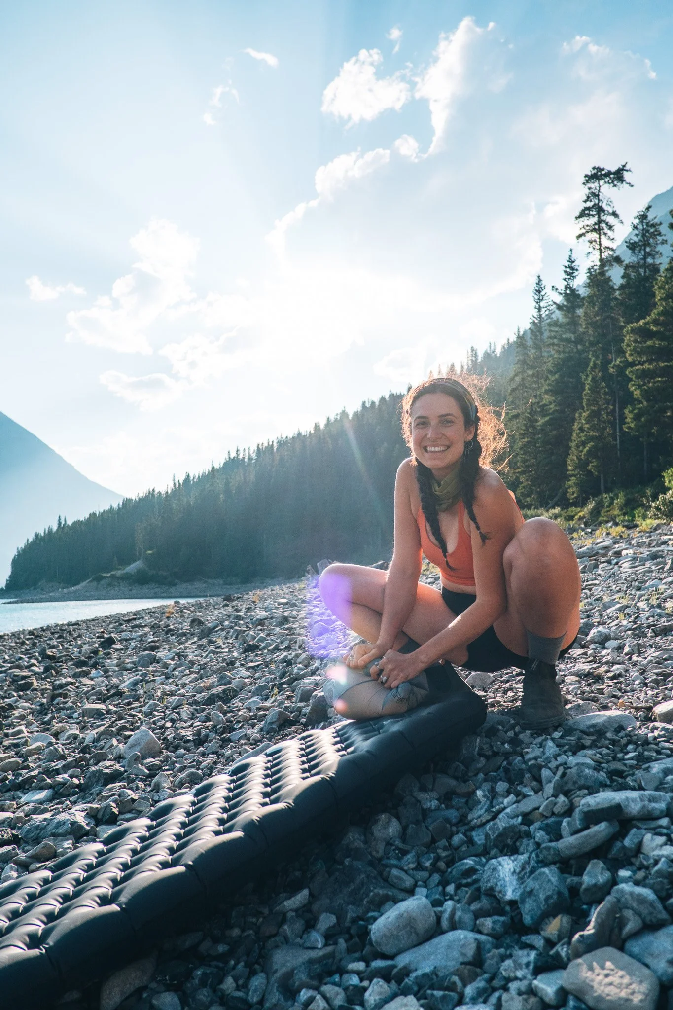 Eine lachende Frau in einem orangefarbenen Tanktop sitzt auf einem felsigen Flussufer und bereitet sich auf eine Wanderung vor, während die Sonne scheint und ein bewaldeter Hügel im Hintergrund sichtbar ist.