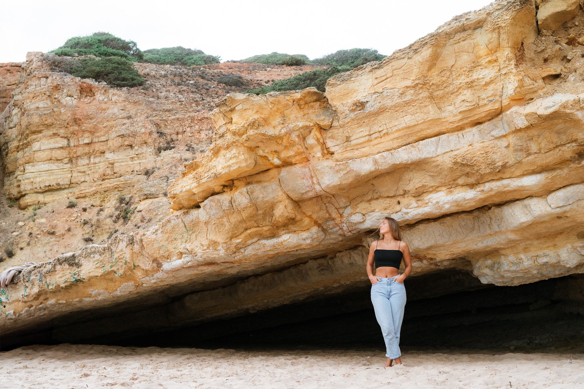 People photography von einer Frau in schwarzem Oberteil und blauen Jeans welche vor einer Felsenhöhle mit großen Felsen im Hintergrund steht.