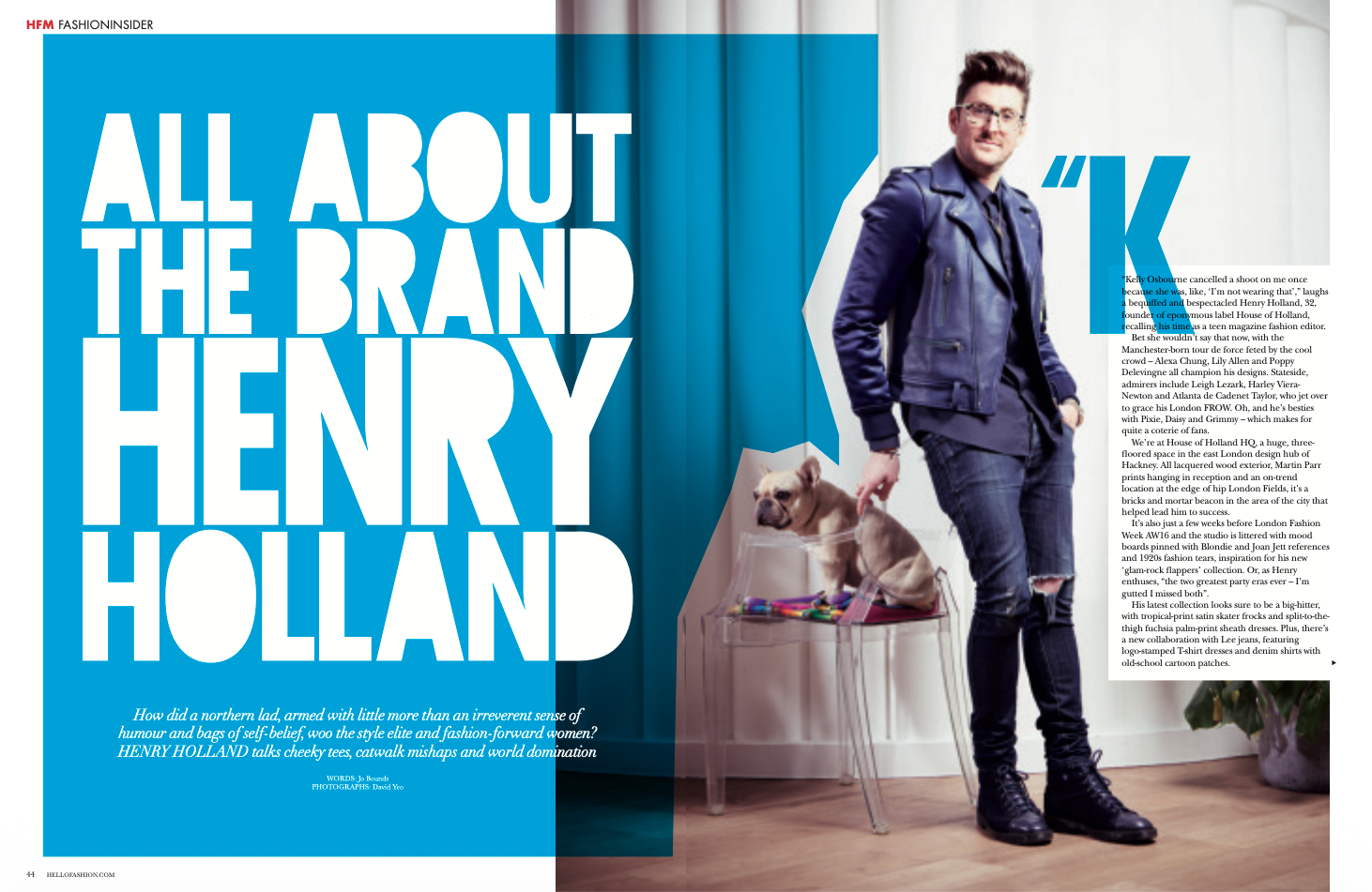 A magazine spread featuring Henry Holland standing indoors, wearing a blue leather jacket, ripped jeans, and black boots. The left page has a bold blue background with large white text that reads 'ALL ABOUT THE BRAND HENRY HOLLAND' and a caption about Henry Holland. The right page contains an article with a small, partial view of a potted plant in the bottom right corner.