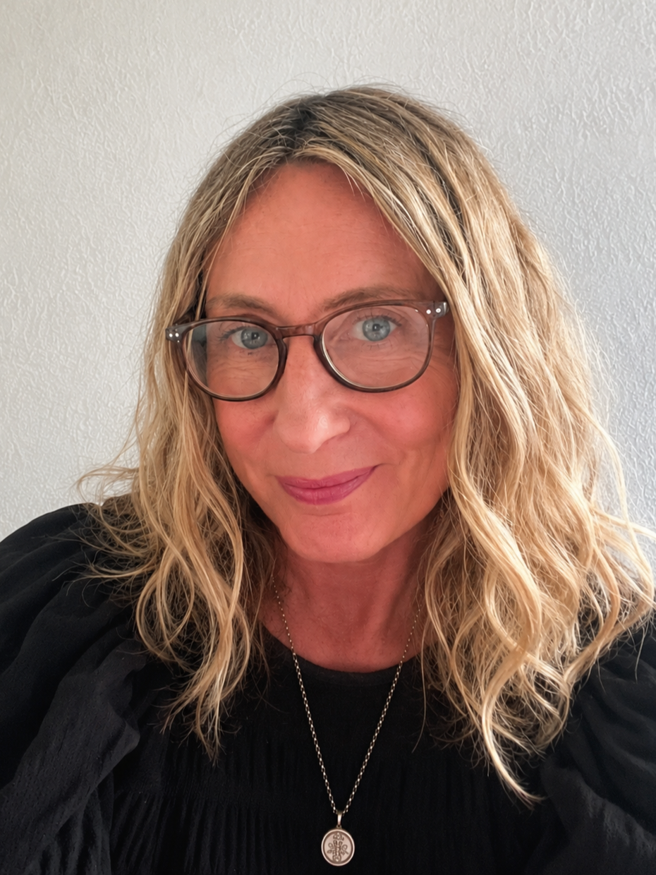 A woman with blonde, curly hair and glasses, smiling, wearing a black top and a necklace, standing against a white textured wall.