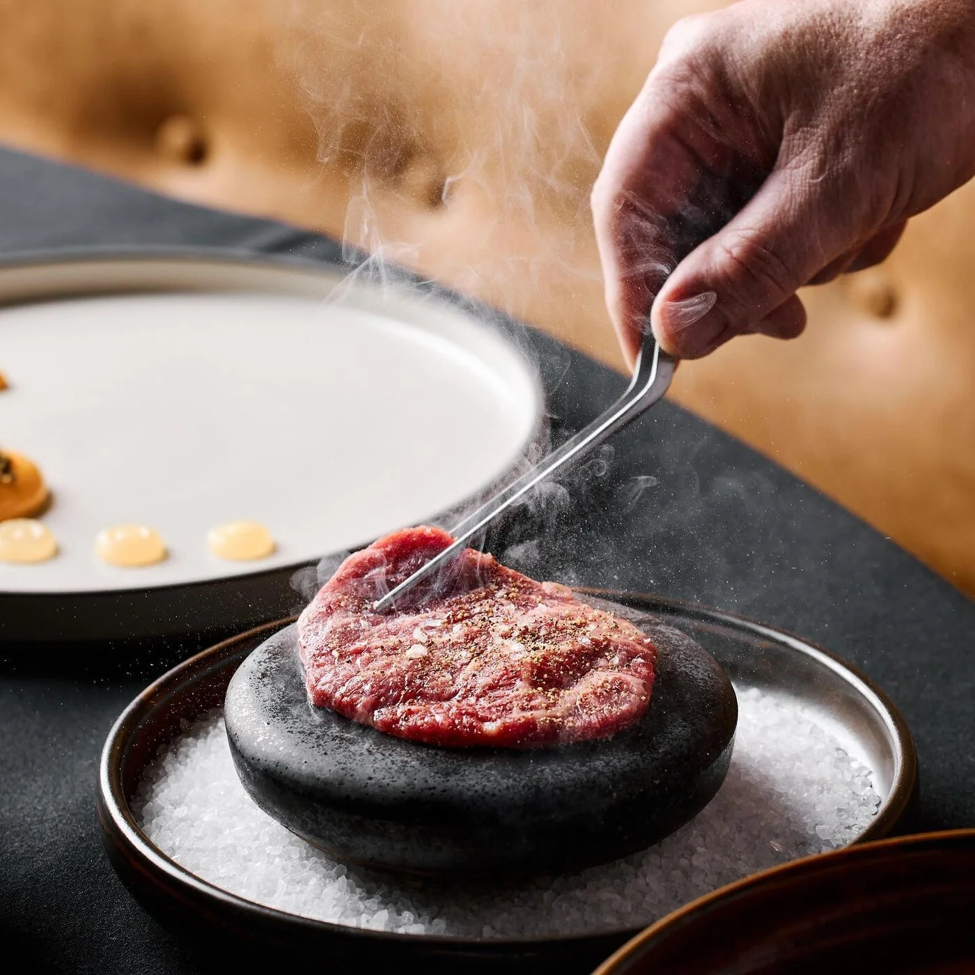 A person using a metal tool to carve into a raw piece of meat on a black stone, with salt around it, and a white plate with small dollops of sauce in the background.