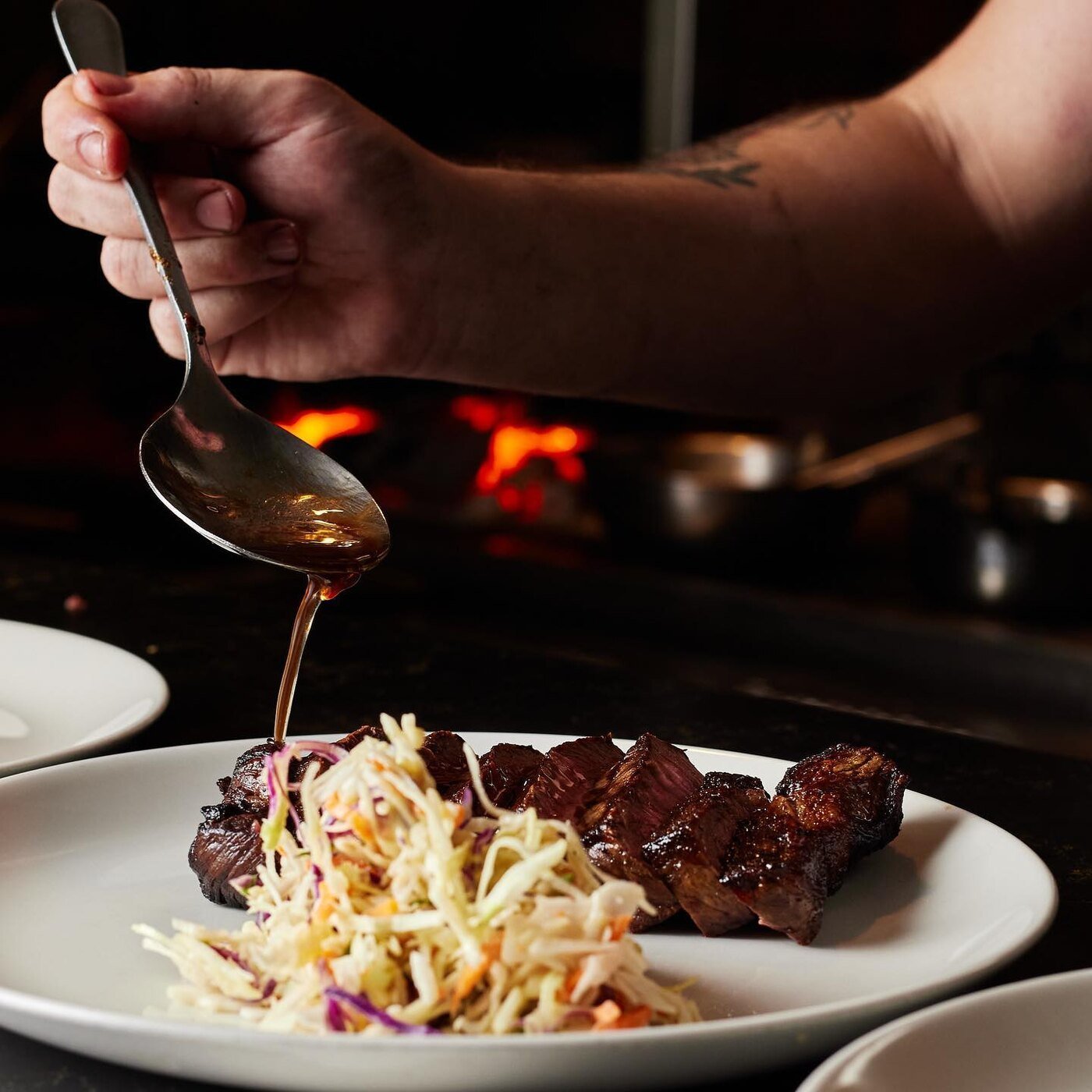 A person pouring sauce over a plate of grilled meat and coleslaw in a restaurant kitchen.