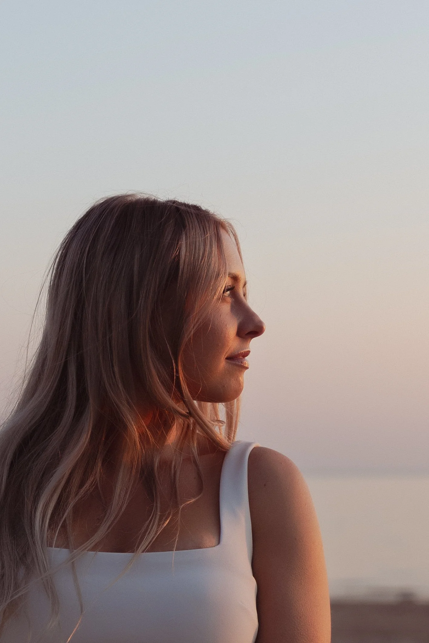 A woman with long, wavy blond hair is in profile, wearing a white sleeveless top, with a calm expression as she looks to the right, with a clear sky background at sunset.