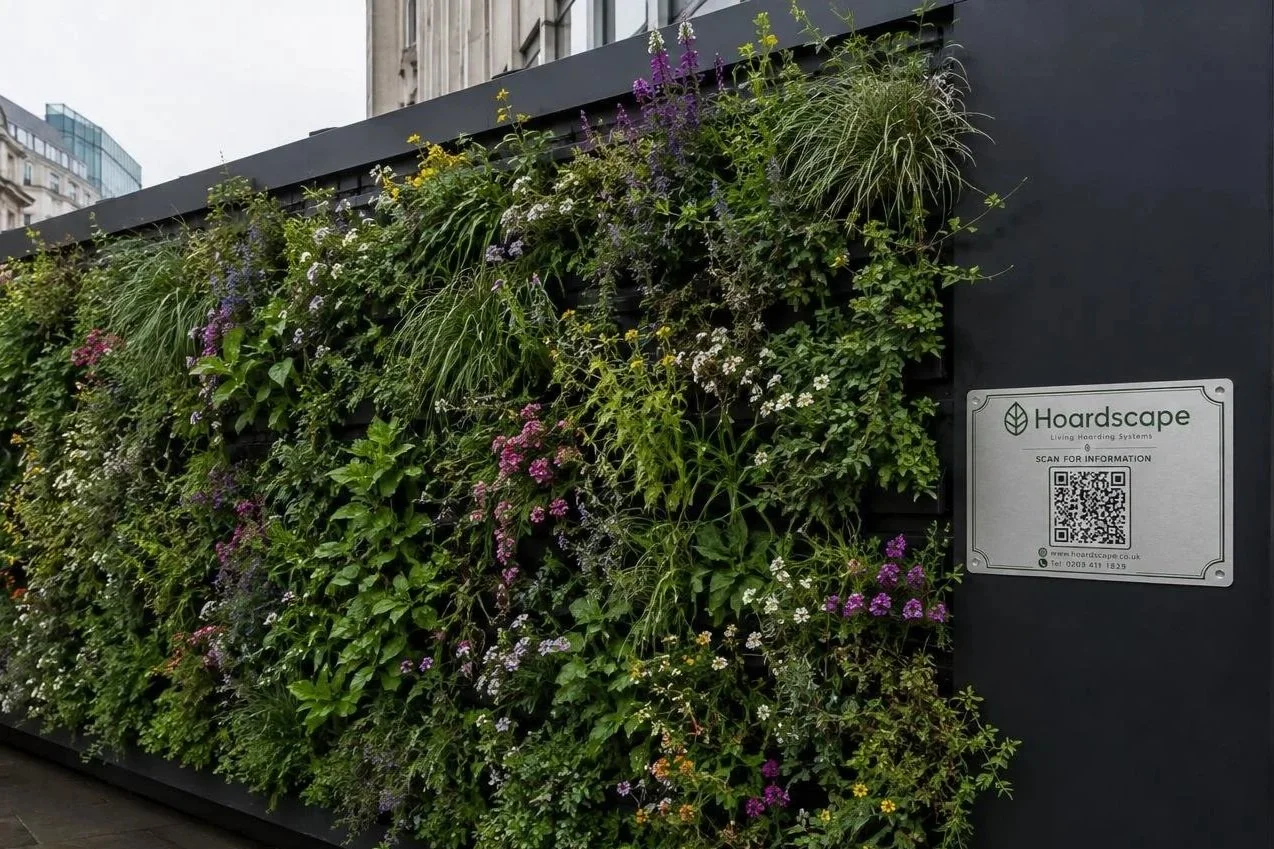 A living green wall with various plants and flowers in front of a gray building, with a sign reading "Hoardscape" and a QR code.