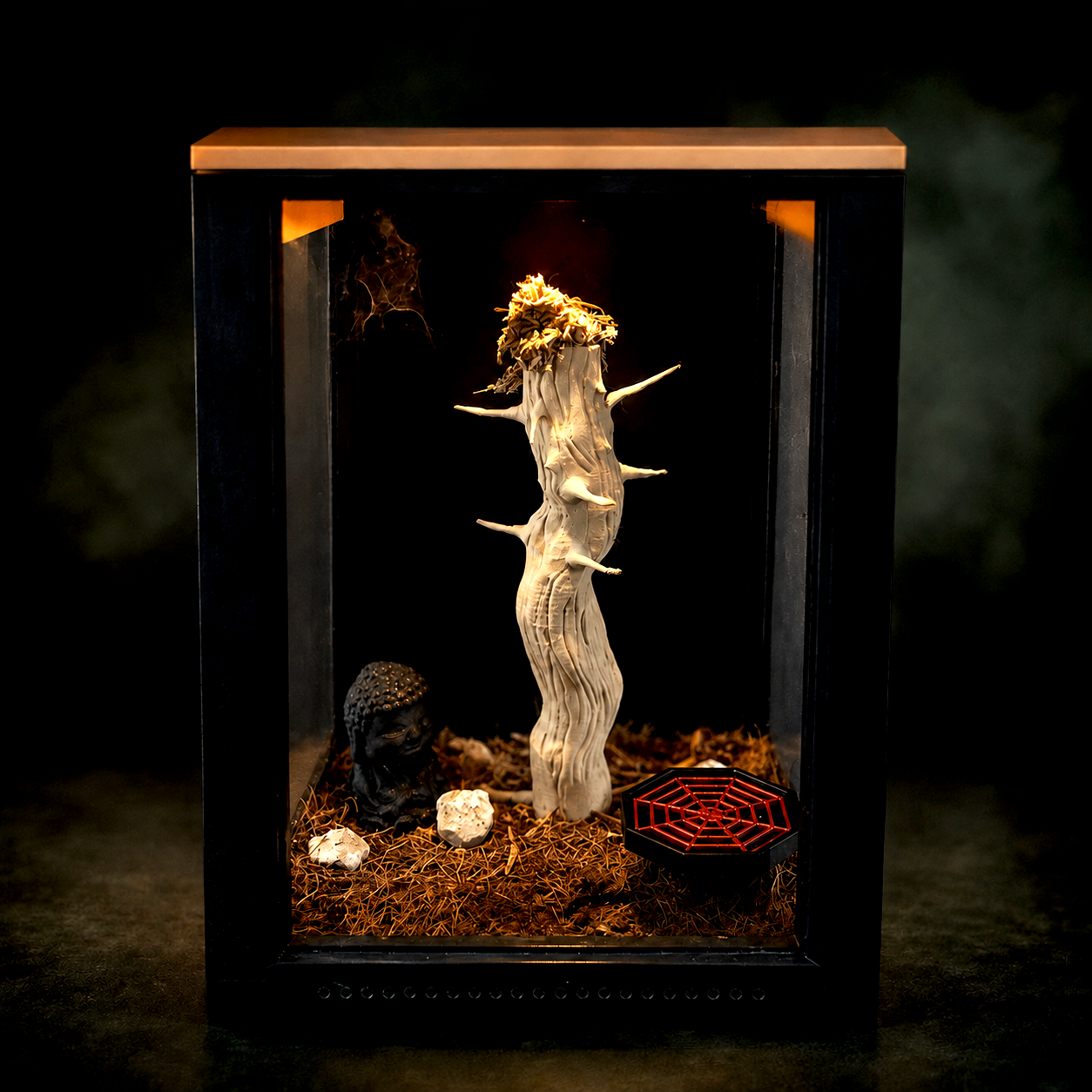 A glass display case with a twisted white tree branch, a small black Buddha head, white rocks, dried grass, and a red spider web decoration inside.