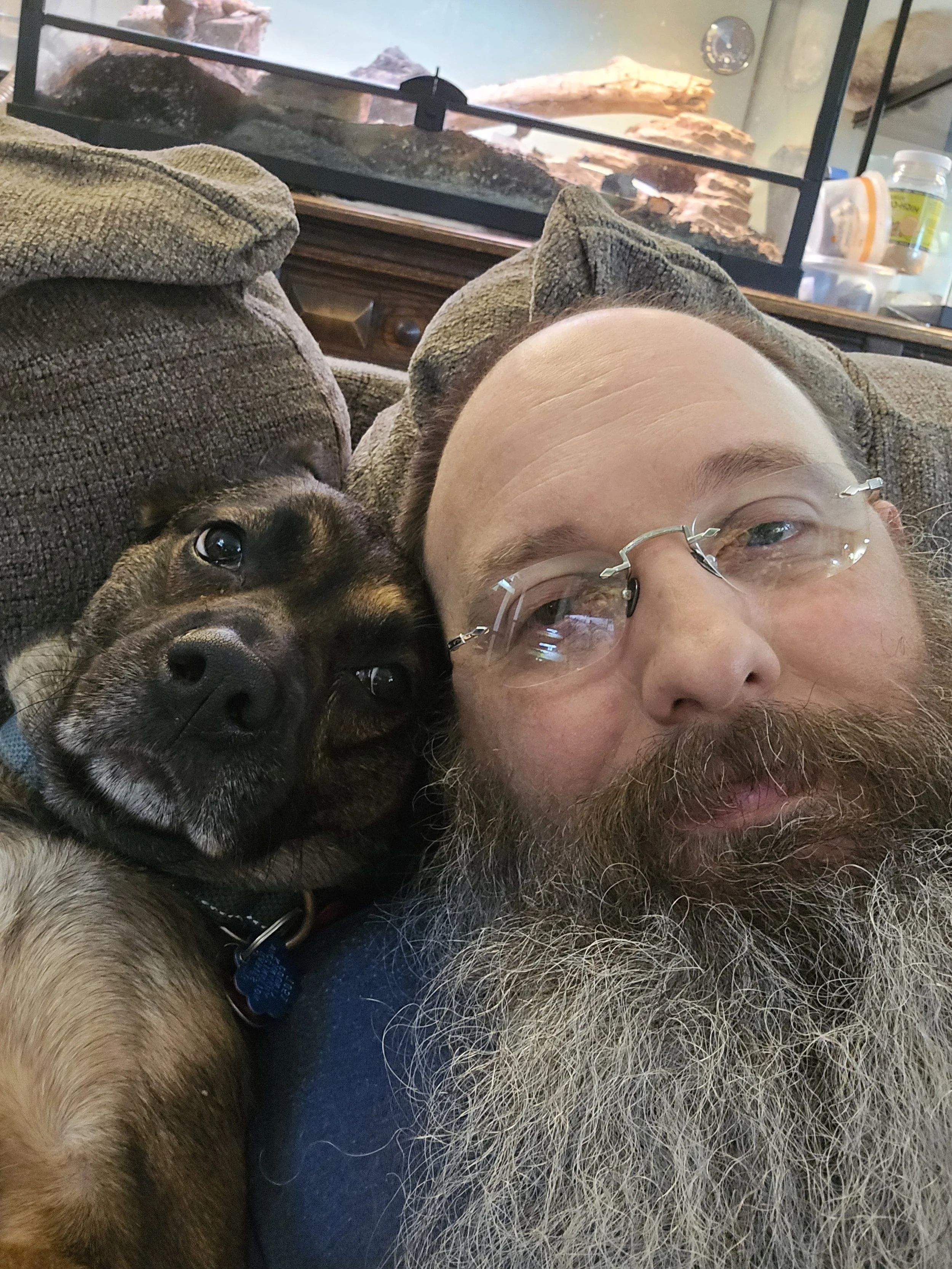 A man with glasses and a beard lying on a couch next to a dog, both looking into the camera, with a terrarium in the background.