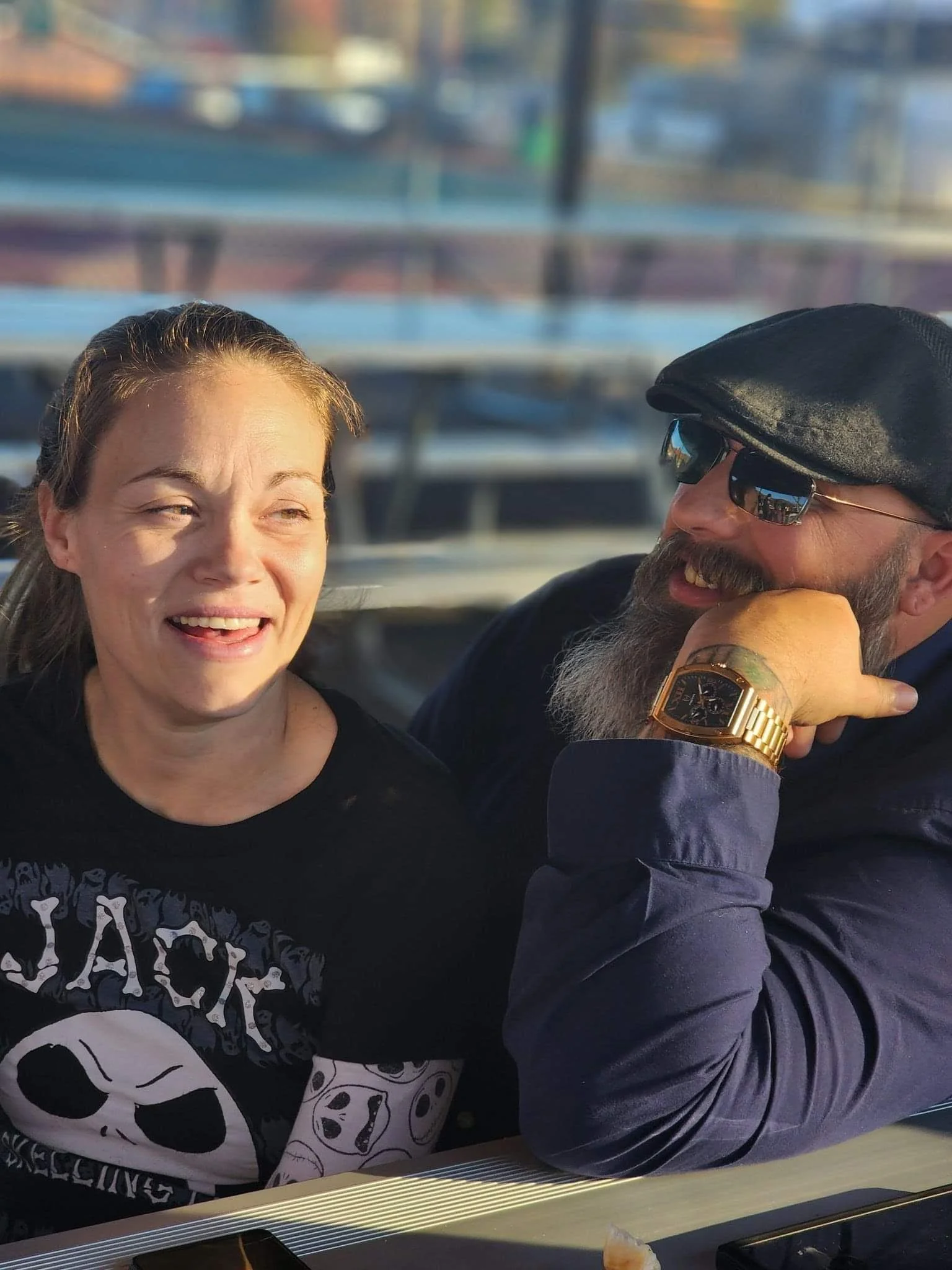 A woman smiling and looking at a man wearing sunglasses and a black beret, who is smiling and resting his chin on his hand. They appear to be enjoying each other's company. The woman has long brown hair, and the man has a beard and is wearing a watch. The background shows blurred outdoor scenery with water and boats.