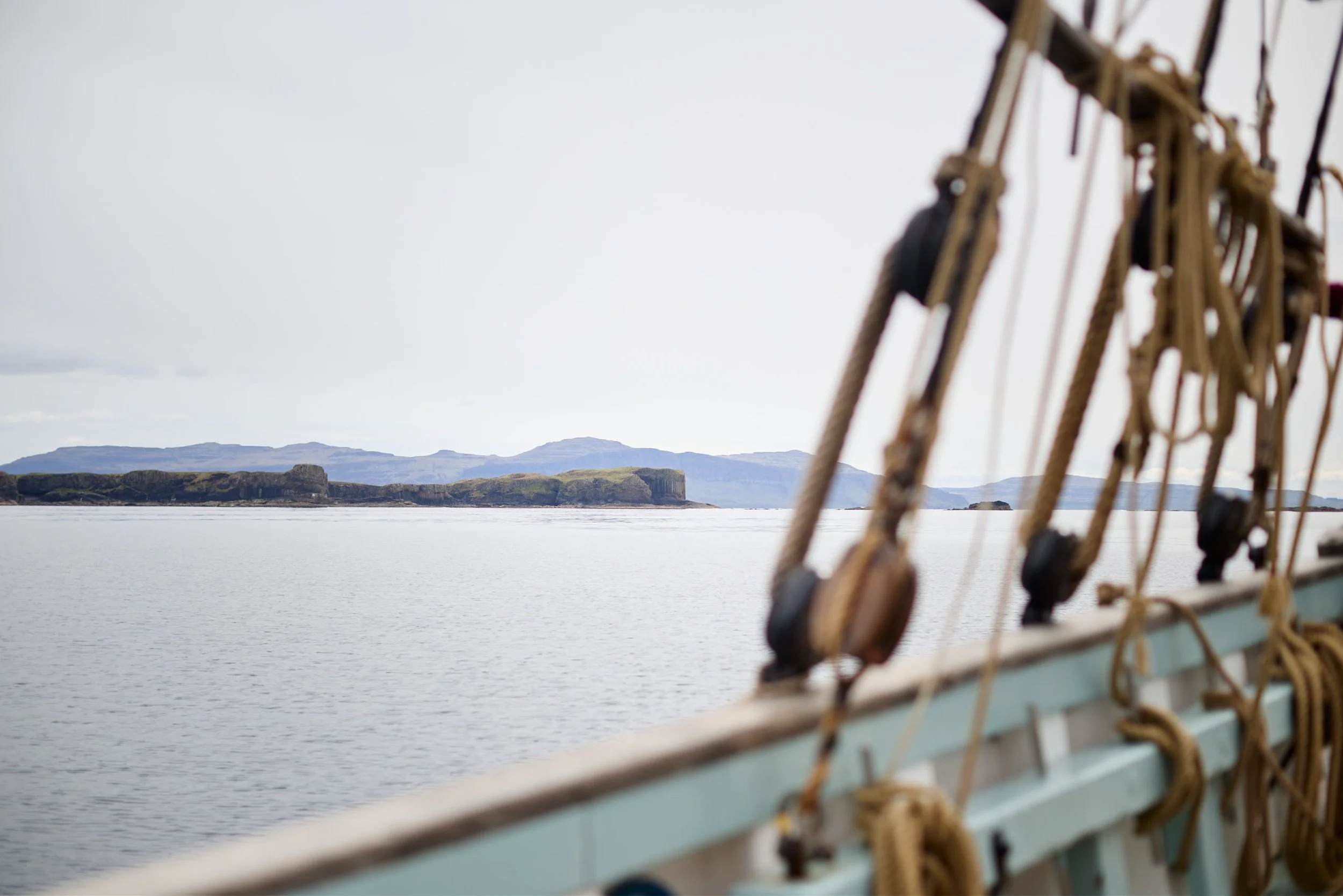 View from a ship's bow showing the ocean with rocky islands and mountainous terrain in the distance, partly obscured by ship rigging.