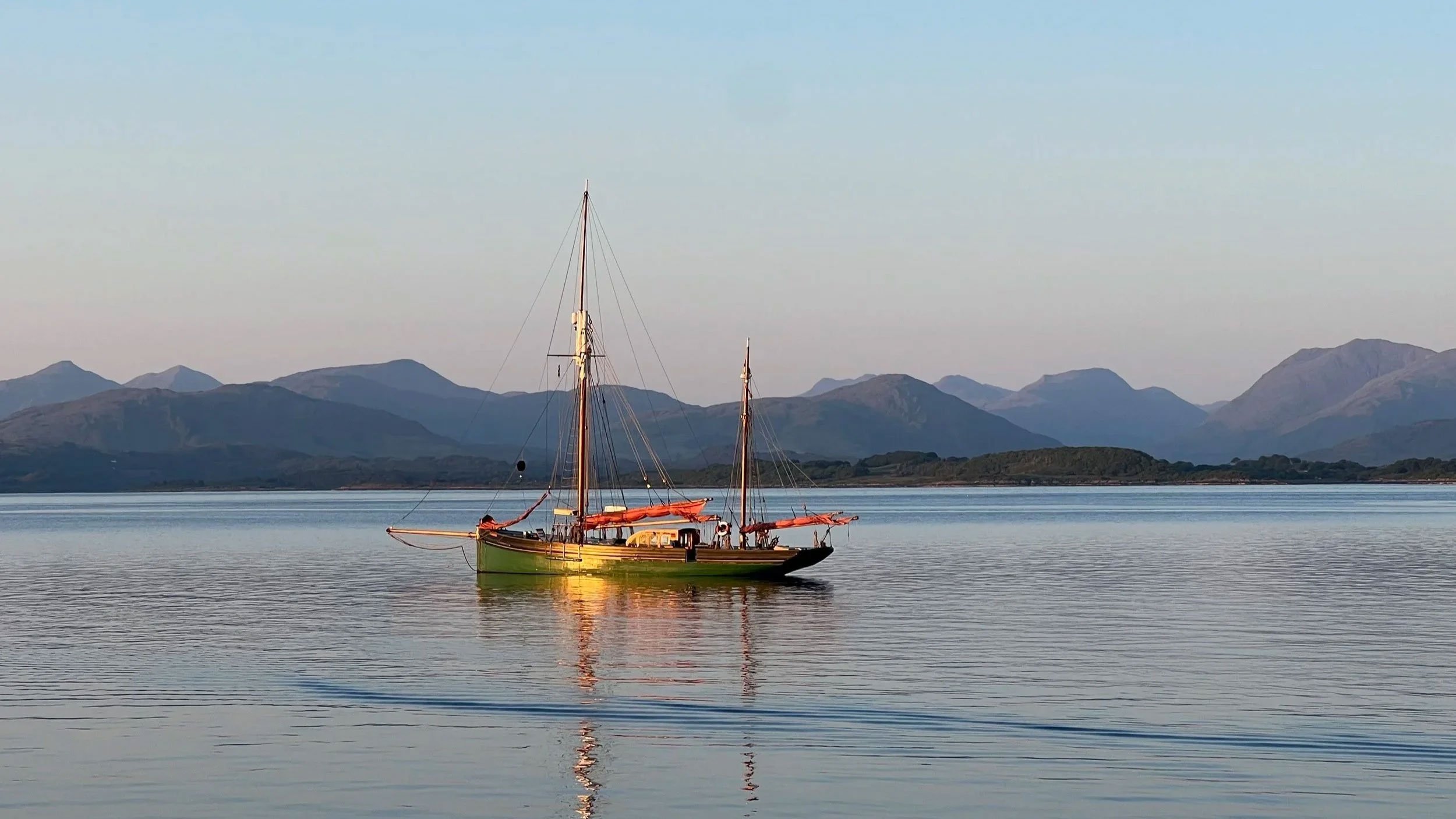 Provident with a green hull floating on calm water with mountain ranges in the background under a clear sky.