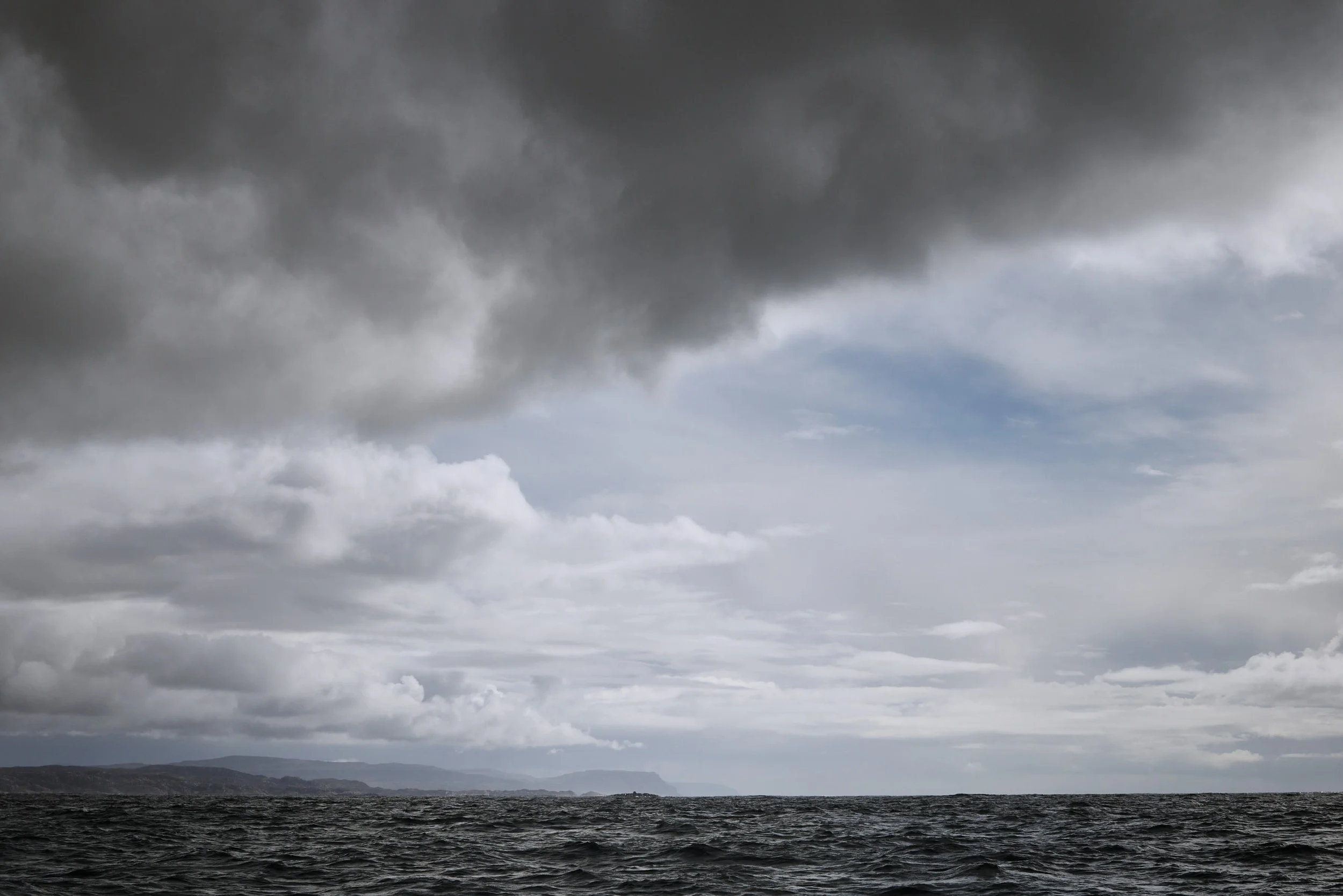 Dark storm clouds over the ocean with distant shoreline in the background.
