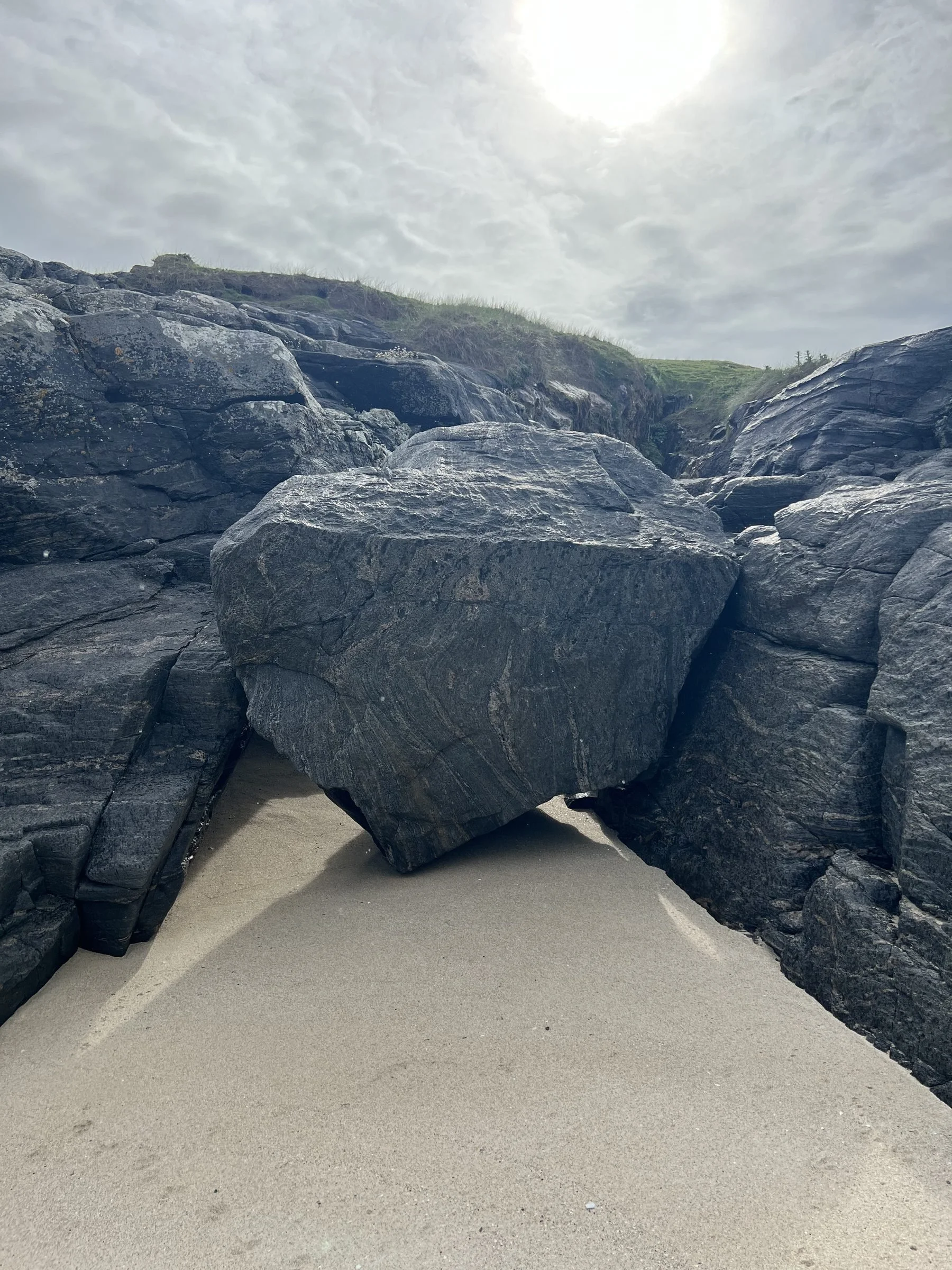Large dark gray rocks on a sandy beach with a cloudy sky and sunlight overhead.