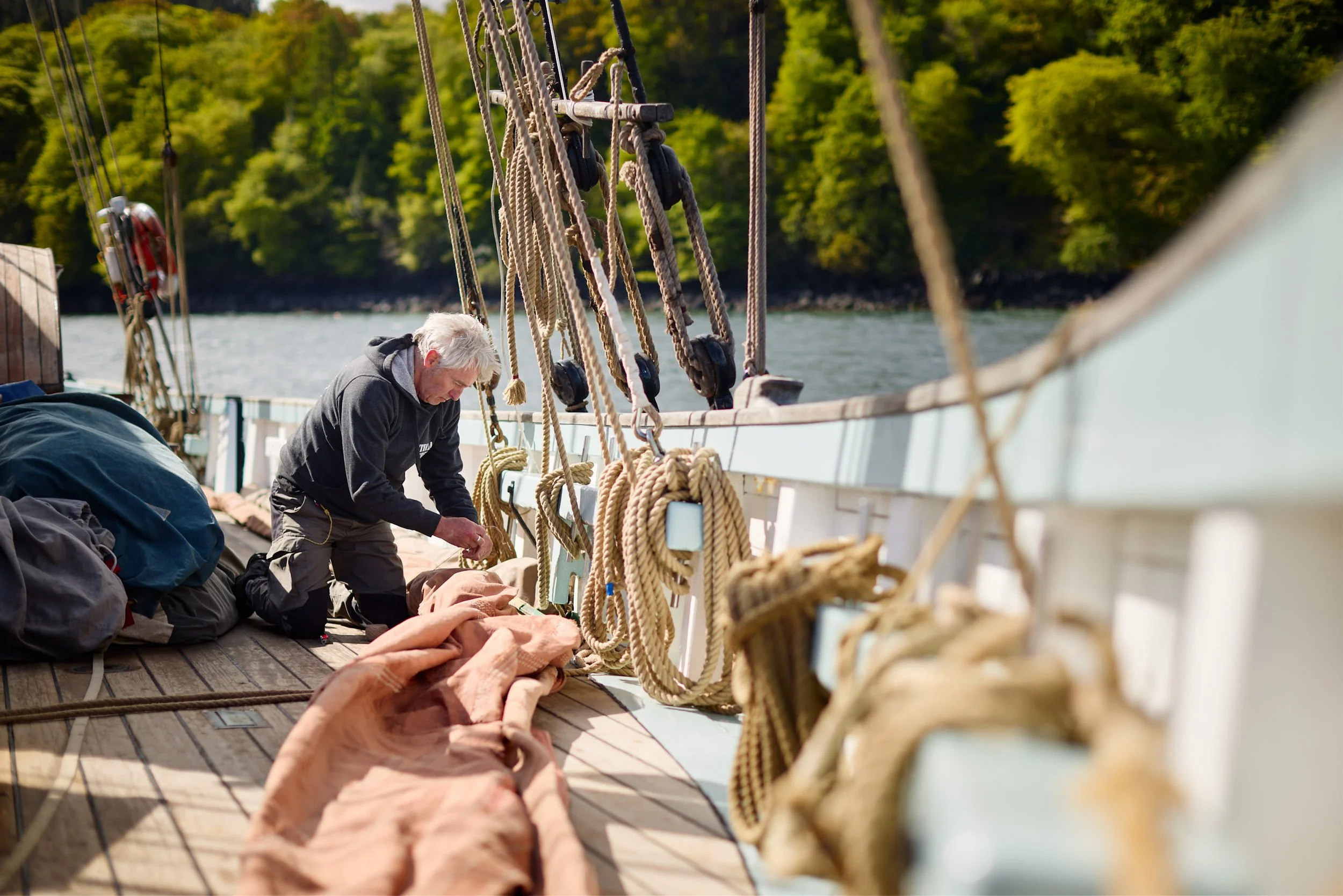 Older man working with ropes and equipment on a sailboat during daytime, with the water and green trees in the background.