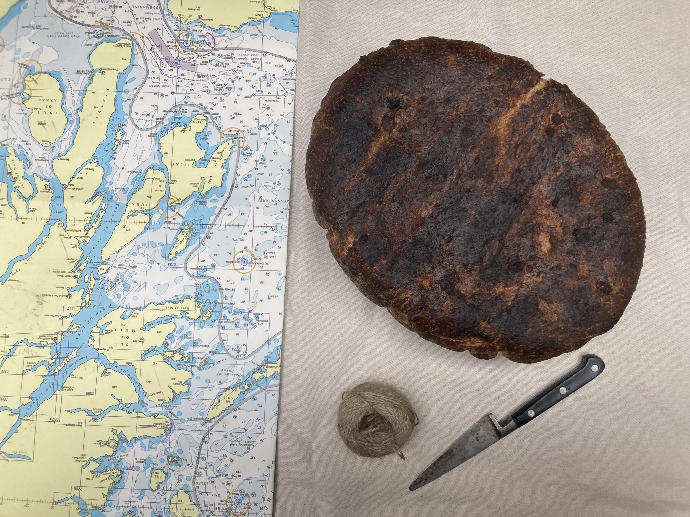 A nautical chart of the Sable Island area on the left side of the image, a large rusty iron rock in the center, a ball of twine below the rock, and a gasket or seal wrench on the bottom right, all arranged on a white cloth surface.