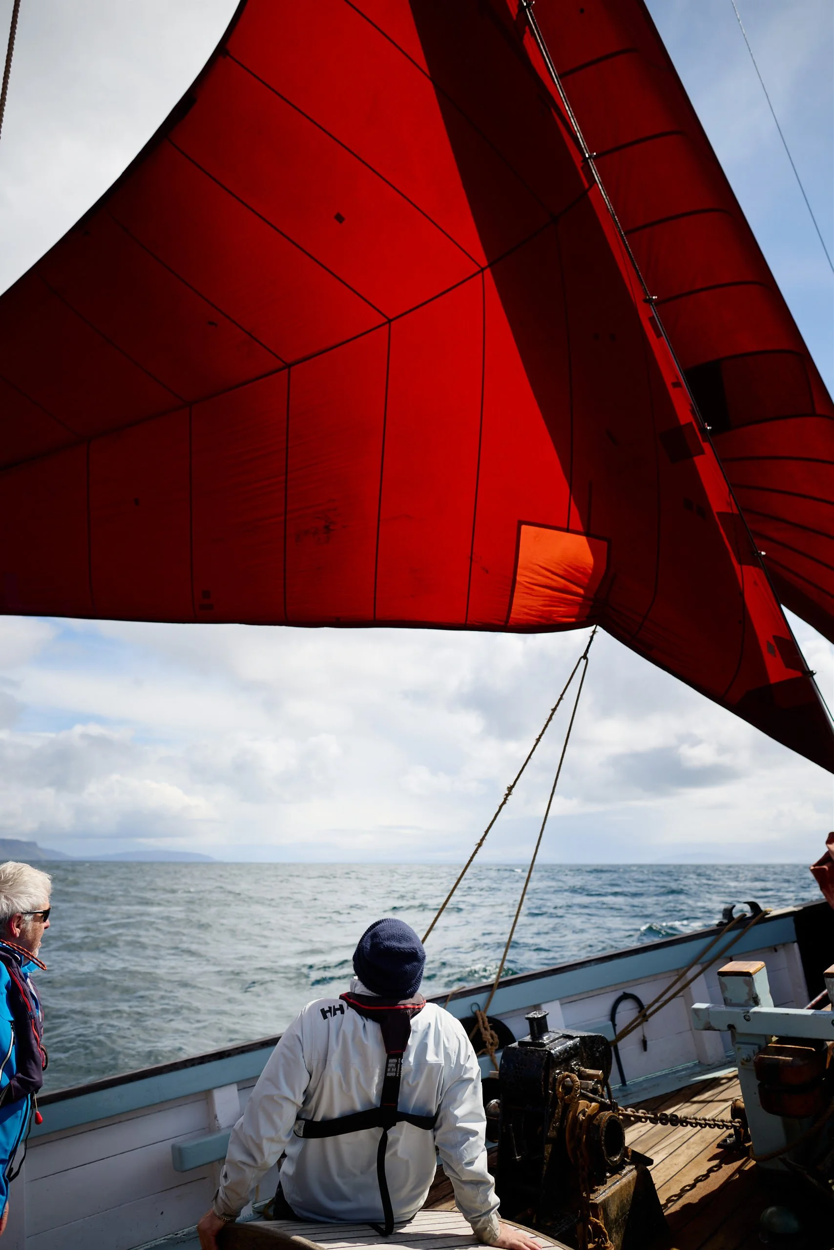 People on Provident racing a sail with a bright red sail against a cloudy sky and ocean background.