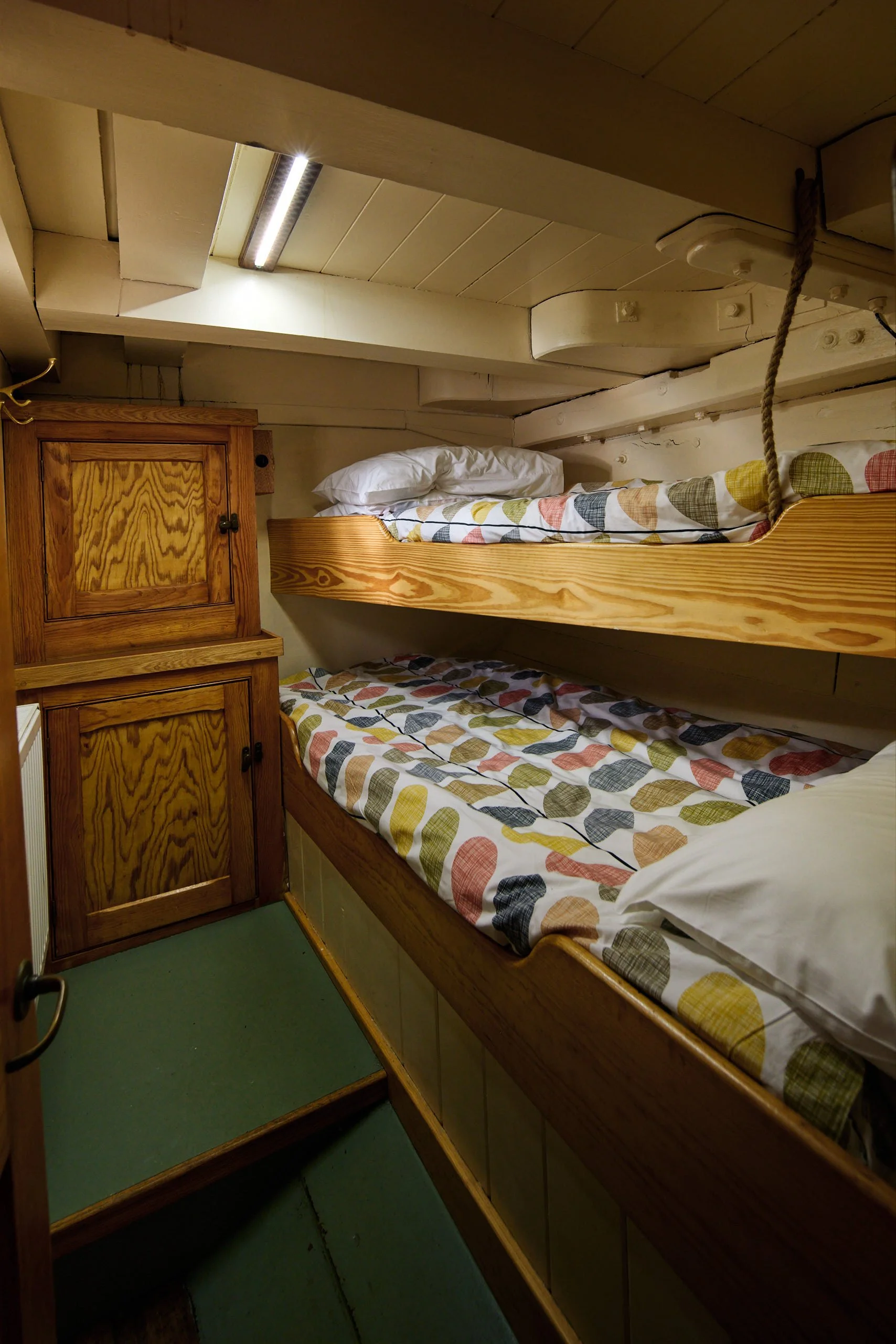 A cozy bedroom with bunk beds. The top bunk has a quilt with a colorful leaf pattern and white pillows. The room is made of wood with a small wooden cabinet and a green floor.