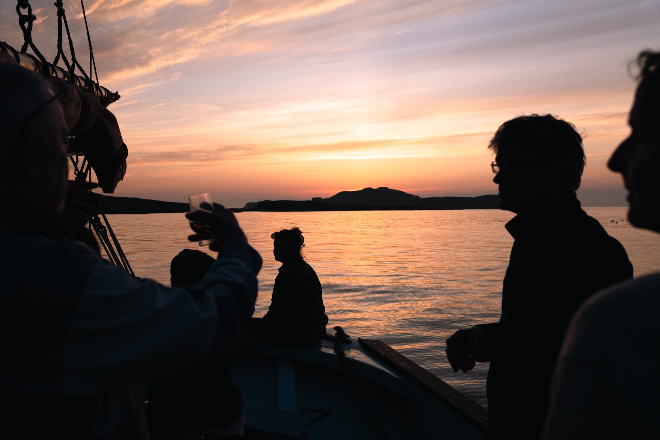 Silhouettes of people on a boat at sunset with a calm body of water and a distant island or landmass in the background.