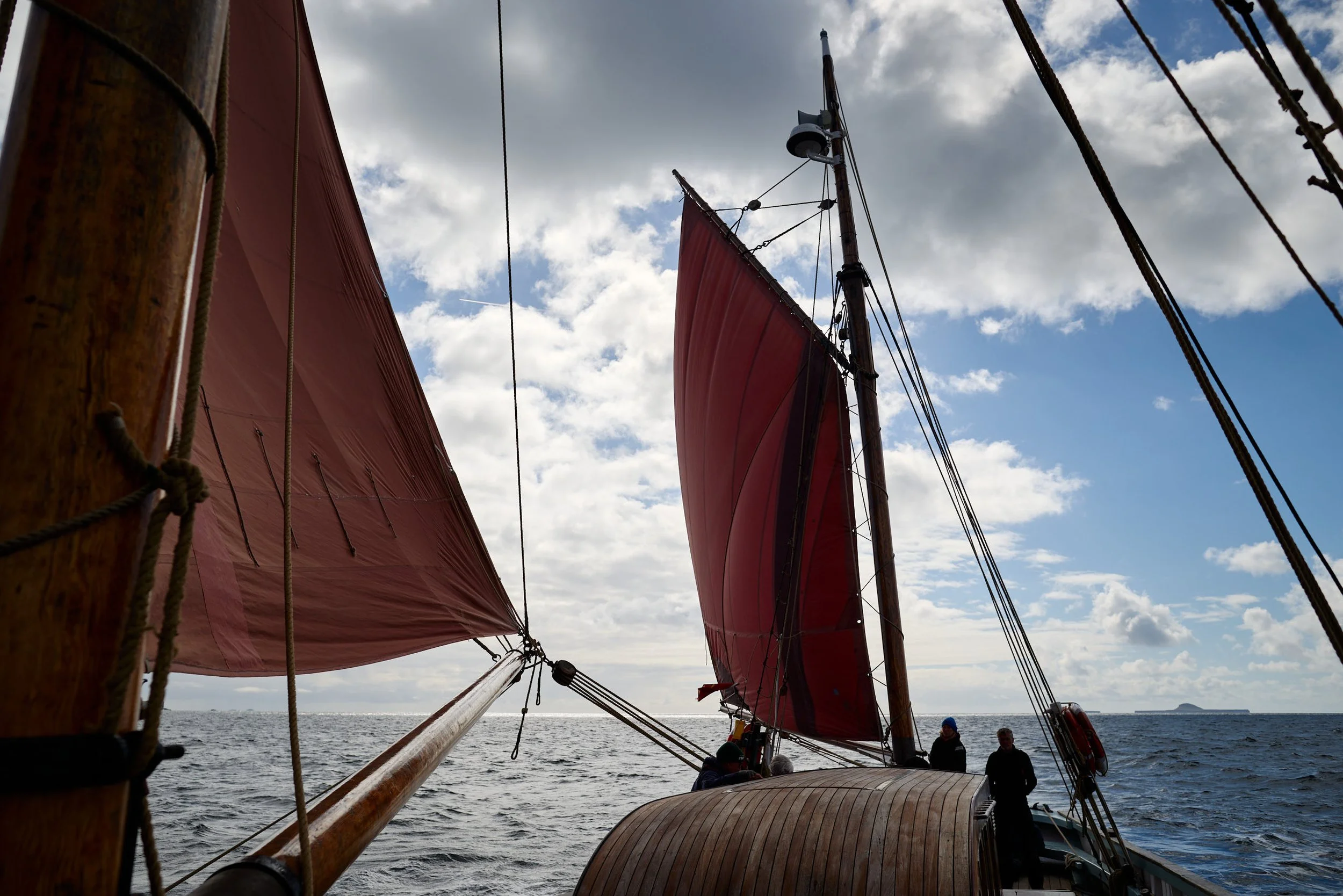 Looking up at a sailboat's masts and red sails against a cloudy sky with the ocean in the background, and a group of people on the deck.