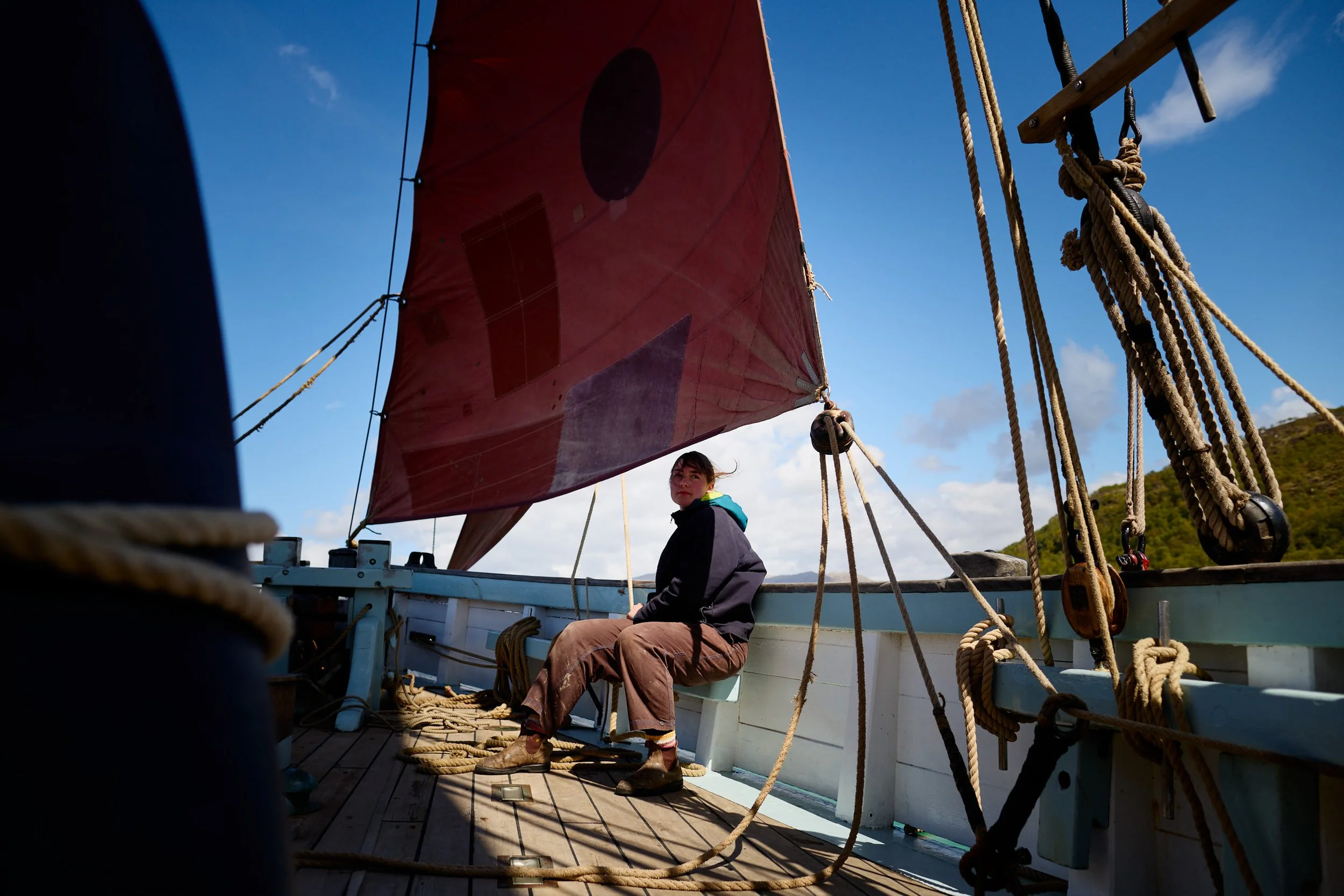 A person sitting on the deck of a sailboat, with a large red sail and numerous ropes, during daylight with a partly cloudy sky.