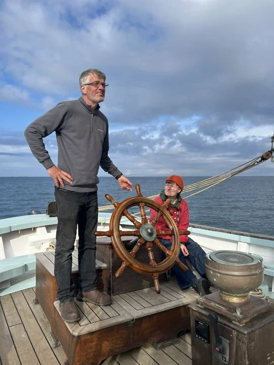 A man standing with his hand on his hip and a woman sitting beside a ship's wheel on a boat at sea under a cloudy sky.
