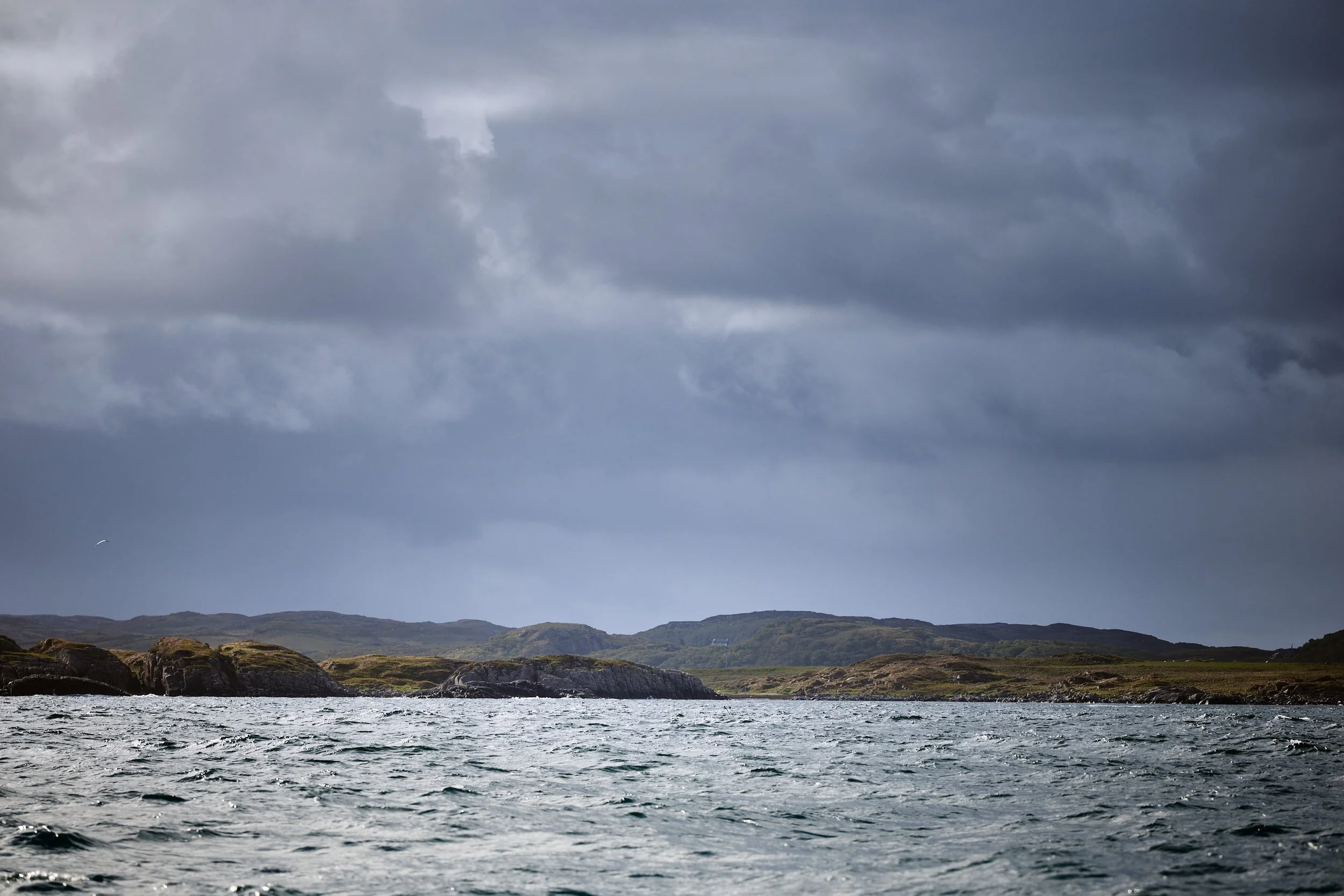 View of a lake with rippling water in the foreground, and rolling green hills under a dark, cloudy sky in the background.
