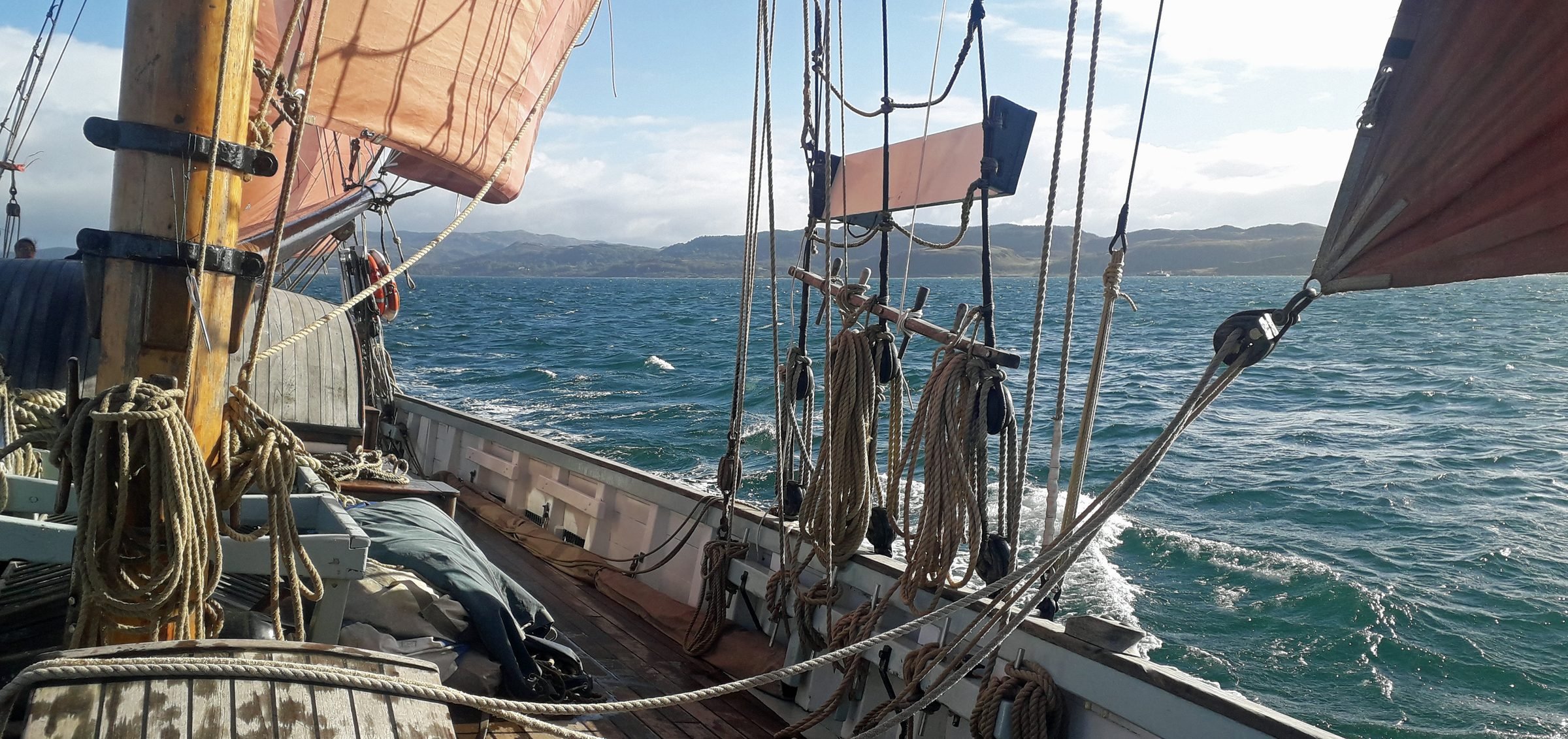 View from a sailboat showing the deck with ropes, sails, and rigging, as well as the open water and distant land with hills in the background.