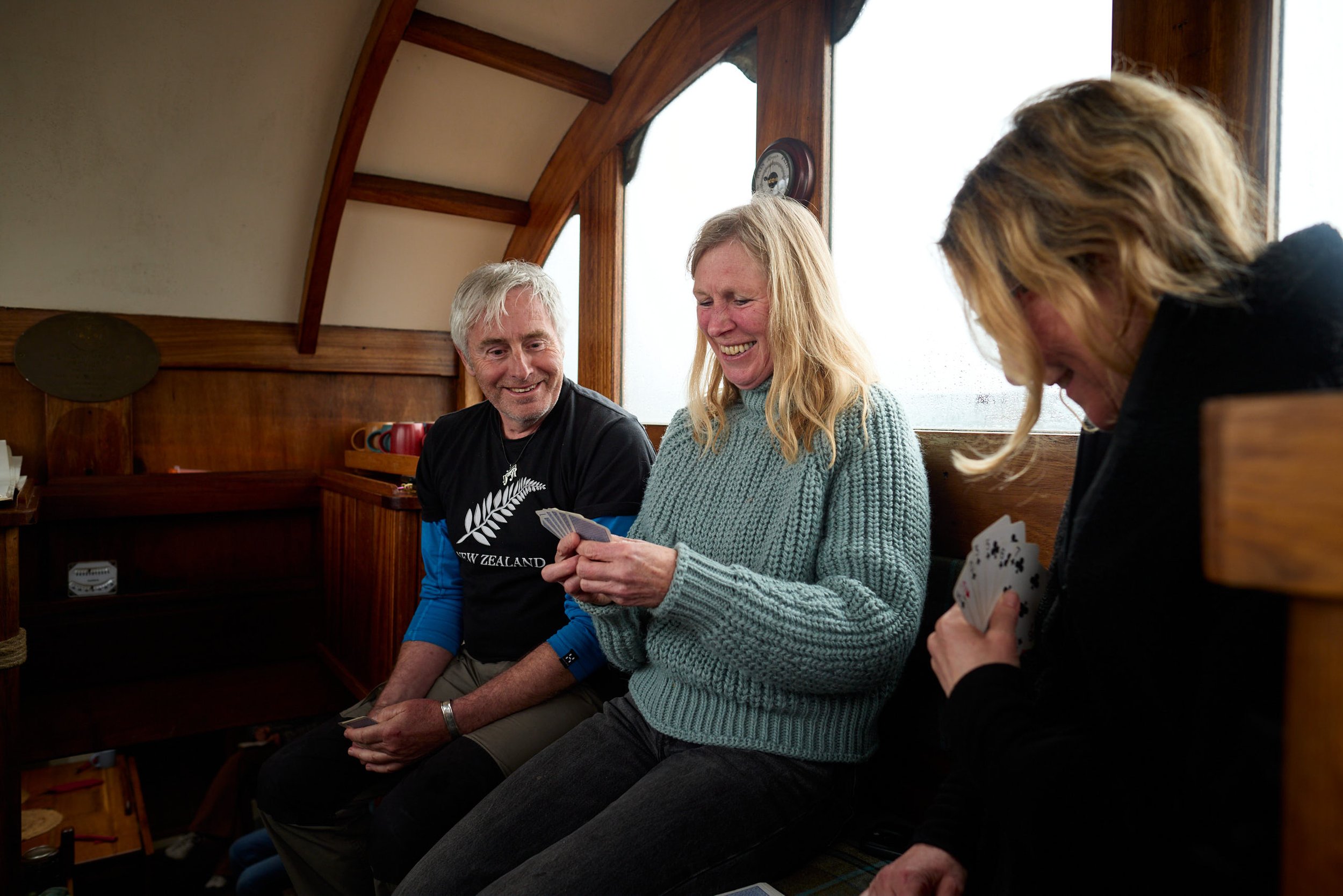 Three people sitting on a wooden bench inside a cozy cabin, engaged in a card game, with smiling faces and a warm atmosphere.