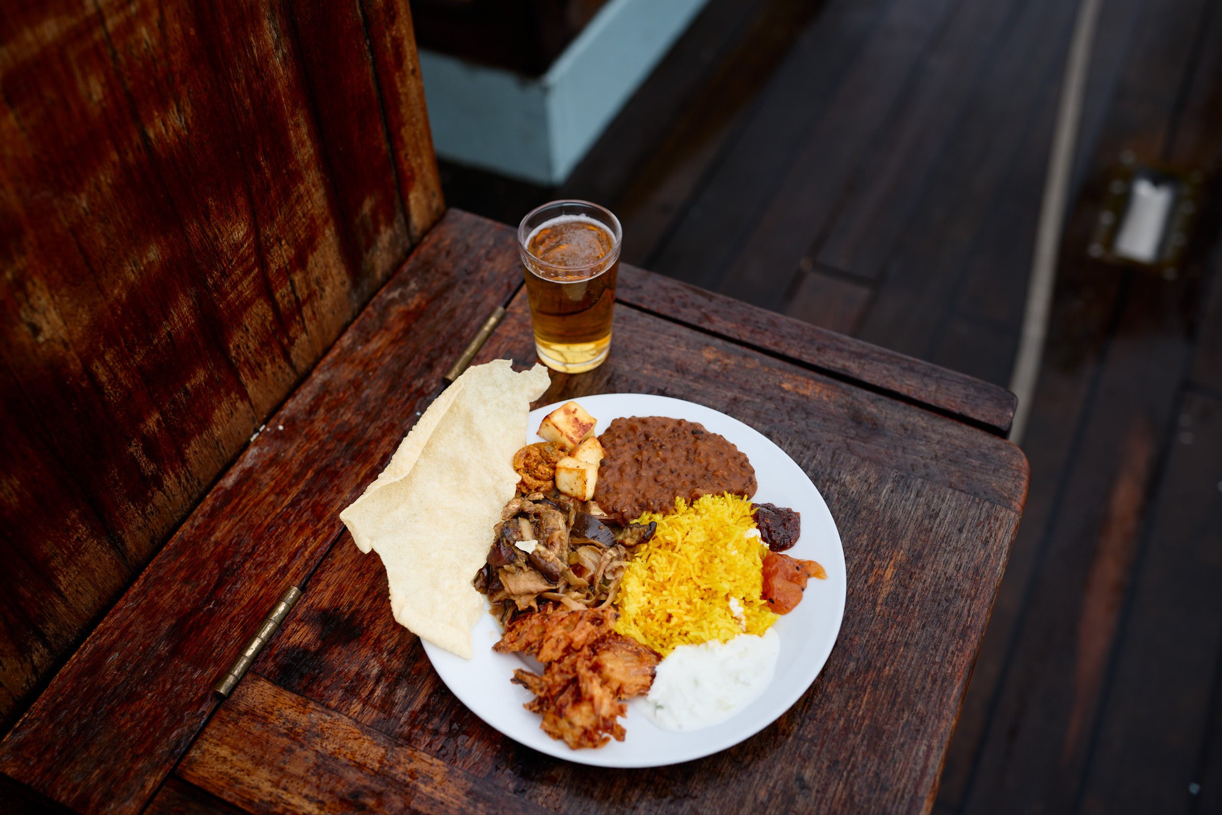 A white plate with Indian food including yellow rice, meat curry, vegetables, papadum, and fried chicken, on a wooden table with a glass of light beer nearby.