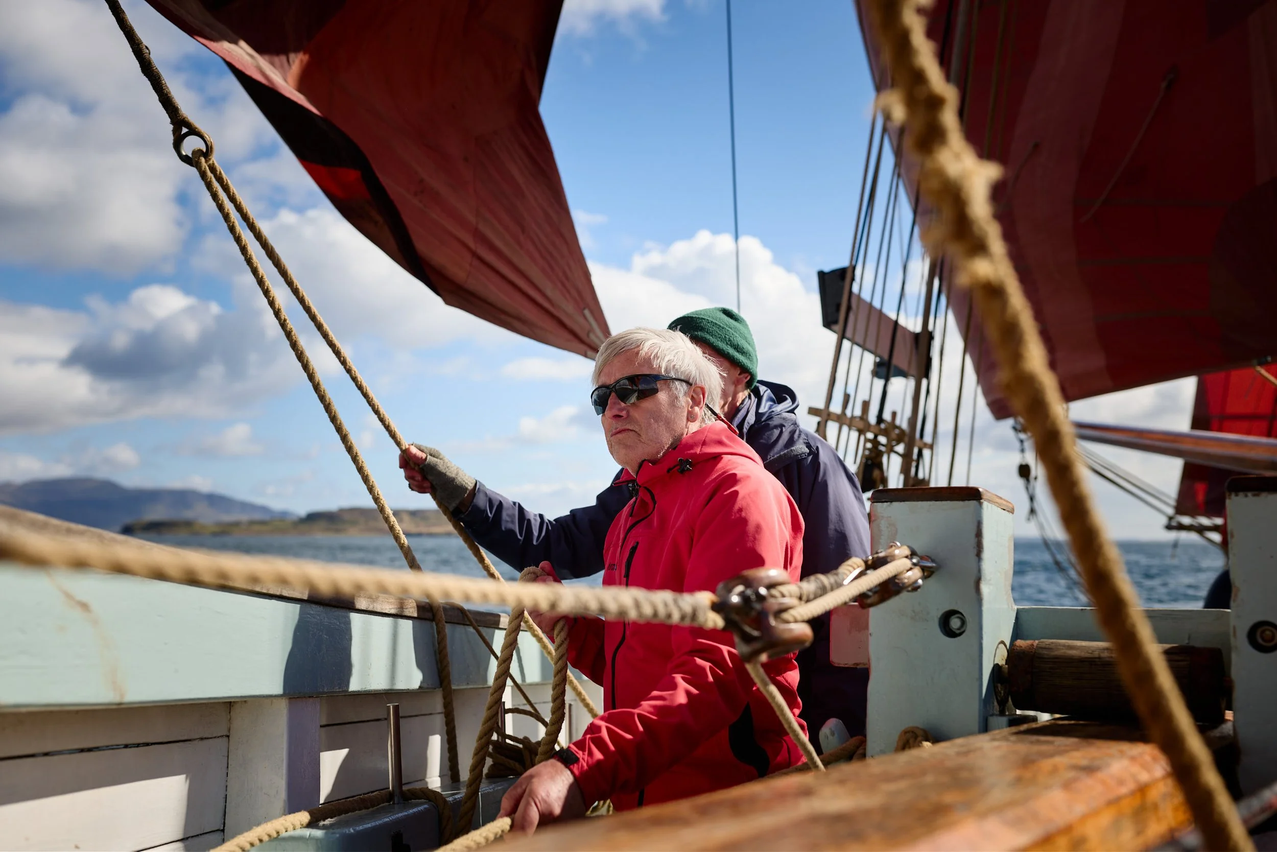 Two men sailing a boat on open water with a landscape in the background under a partly cloudy sky, one man in a red jacket and sunglasses.
