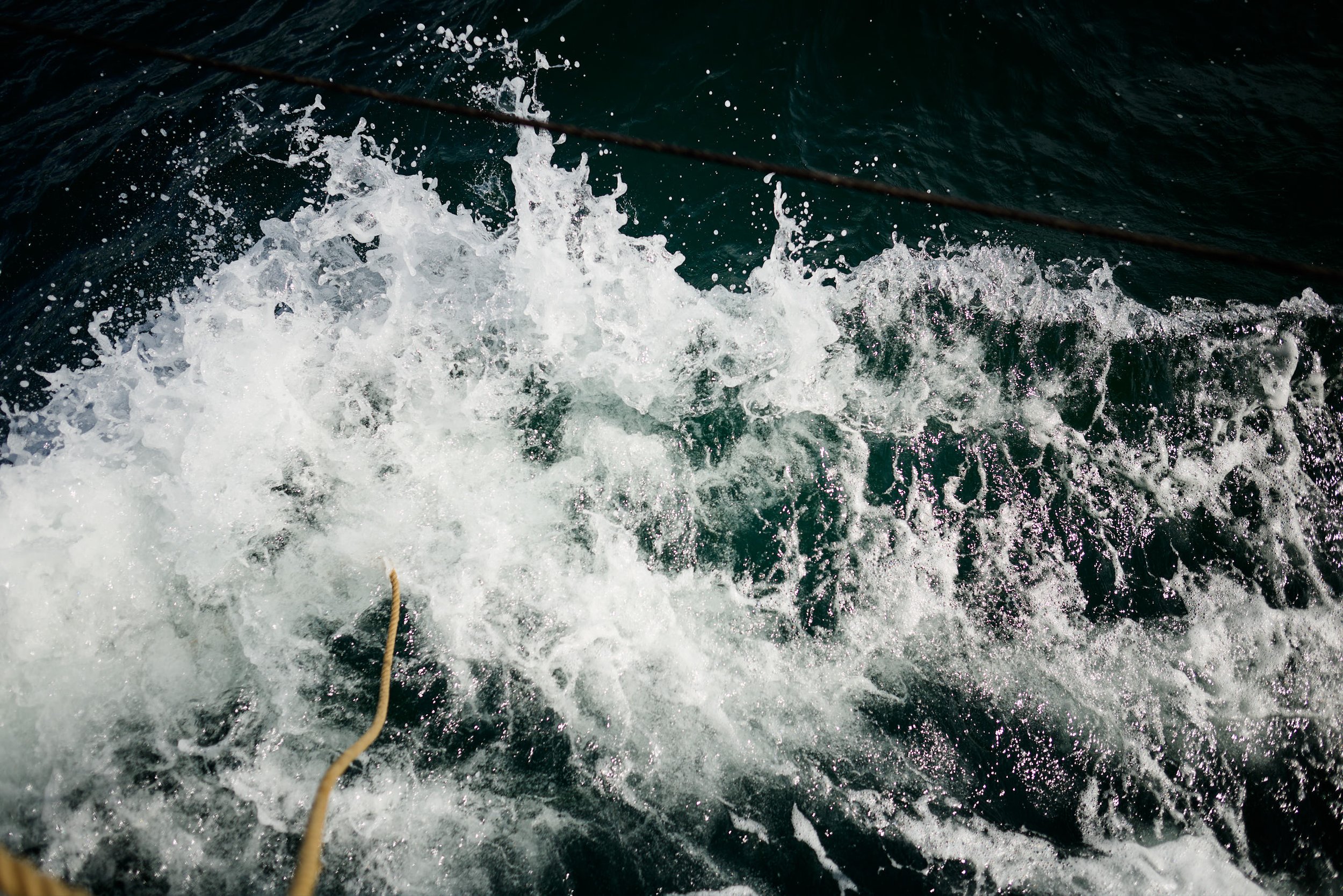 View of water splashing behind a boat, with a rope in the foreground and a diagonal line across the image likely from the boat's structure.