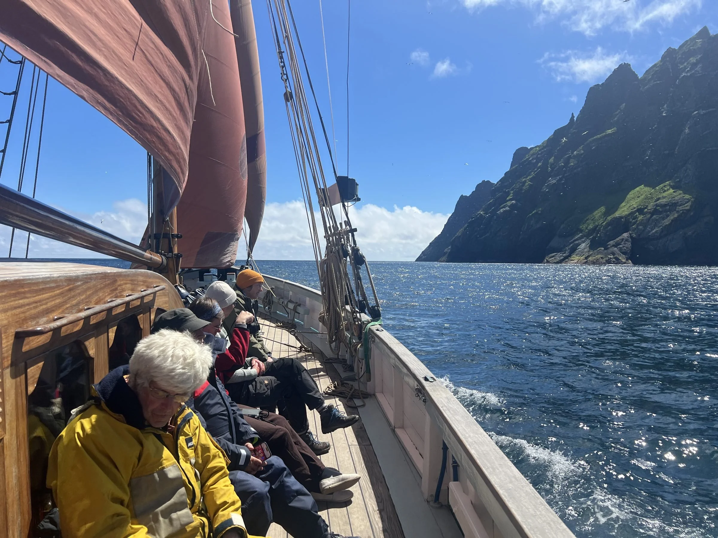 Passengers sitting on the deck of a sailboat, with a view of the ocean and a steep rocky green mountain in the background under a partly cloudy blue sky.