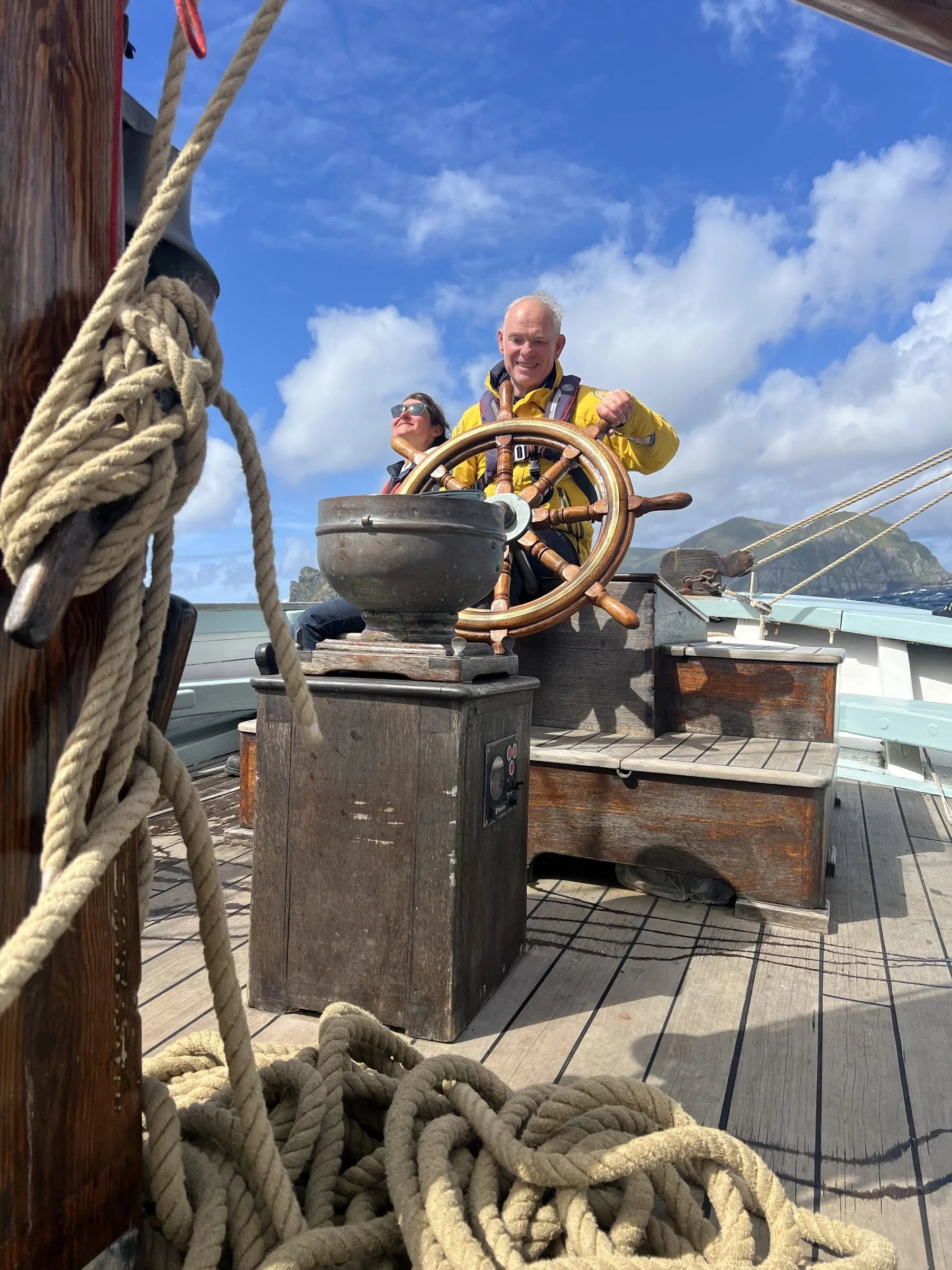 A man at the helm of a boat, smiling, with a woman behind him, on a sunny day with blue sky and clouds, surrounded by a large pile of ropes, wooden deck, and mountain view in the background.