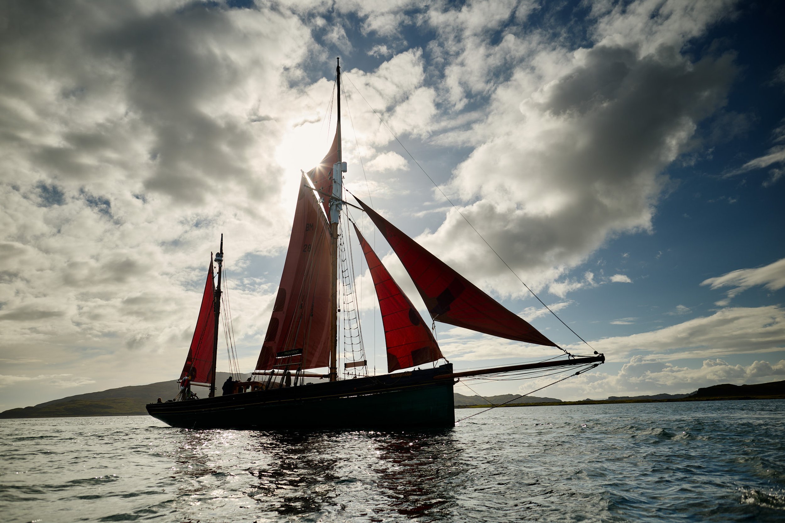 A sailboat with red sails on a body of water, with mountains in the background and a cloudy sky overhead.