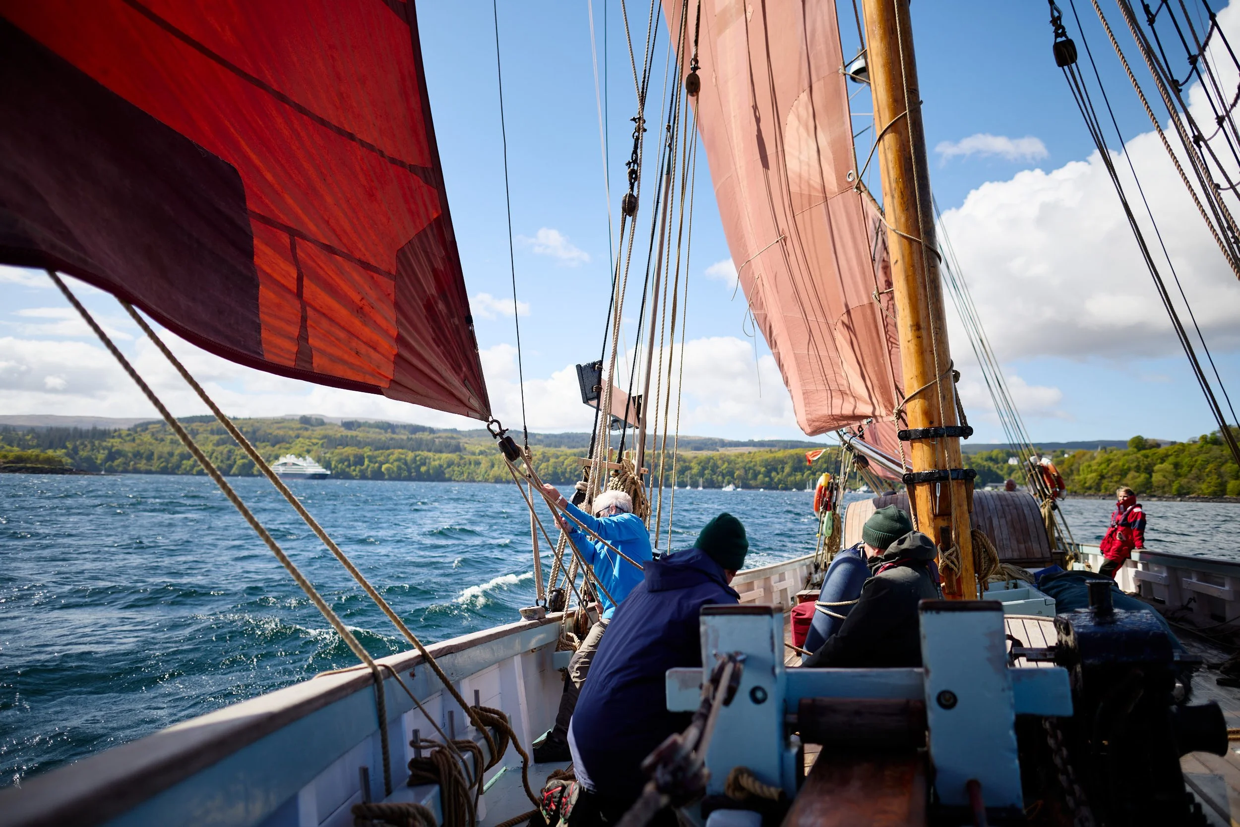 People aboard a sailboat with large red and pink sails on a body of water, with a cruise ship and a green shoreline with trees in the background. The sky is partly cloudy.