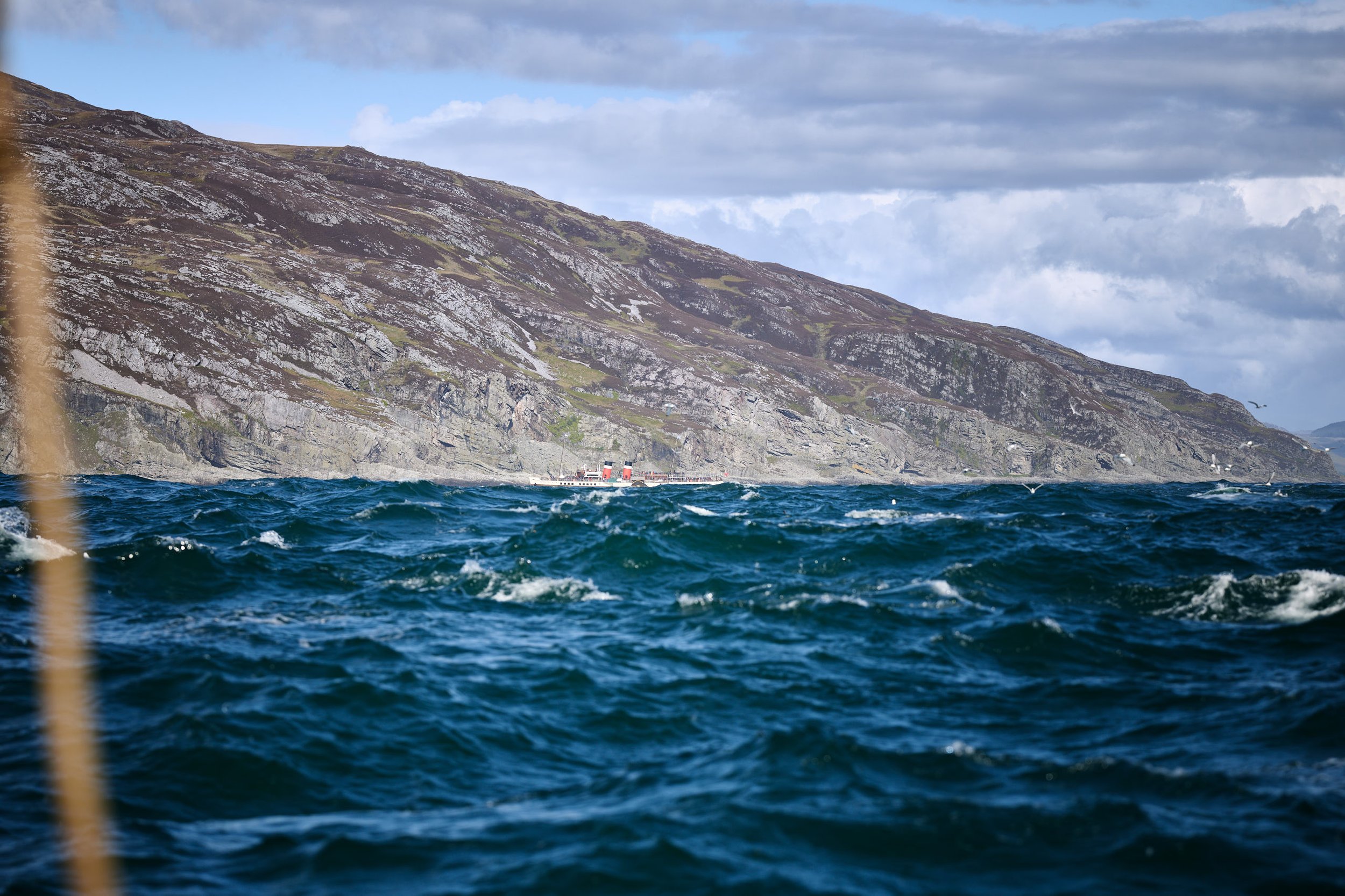 View of a large ship sailing on dark blue ocean with rocky, green-covered mountains and a cloudy sky in the background.