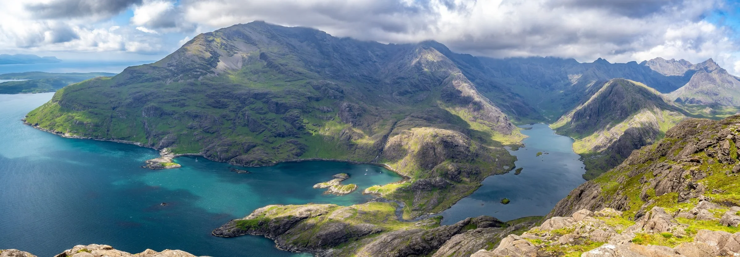Scenic view of a mountain range with green slopes, a lake, and a cloudy sky.