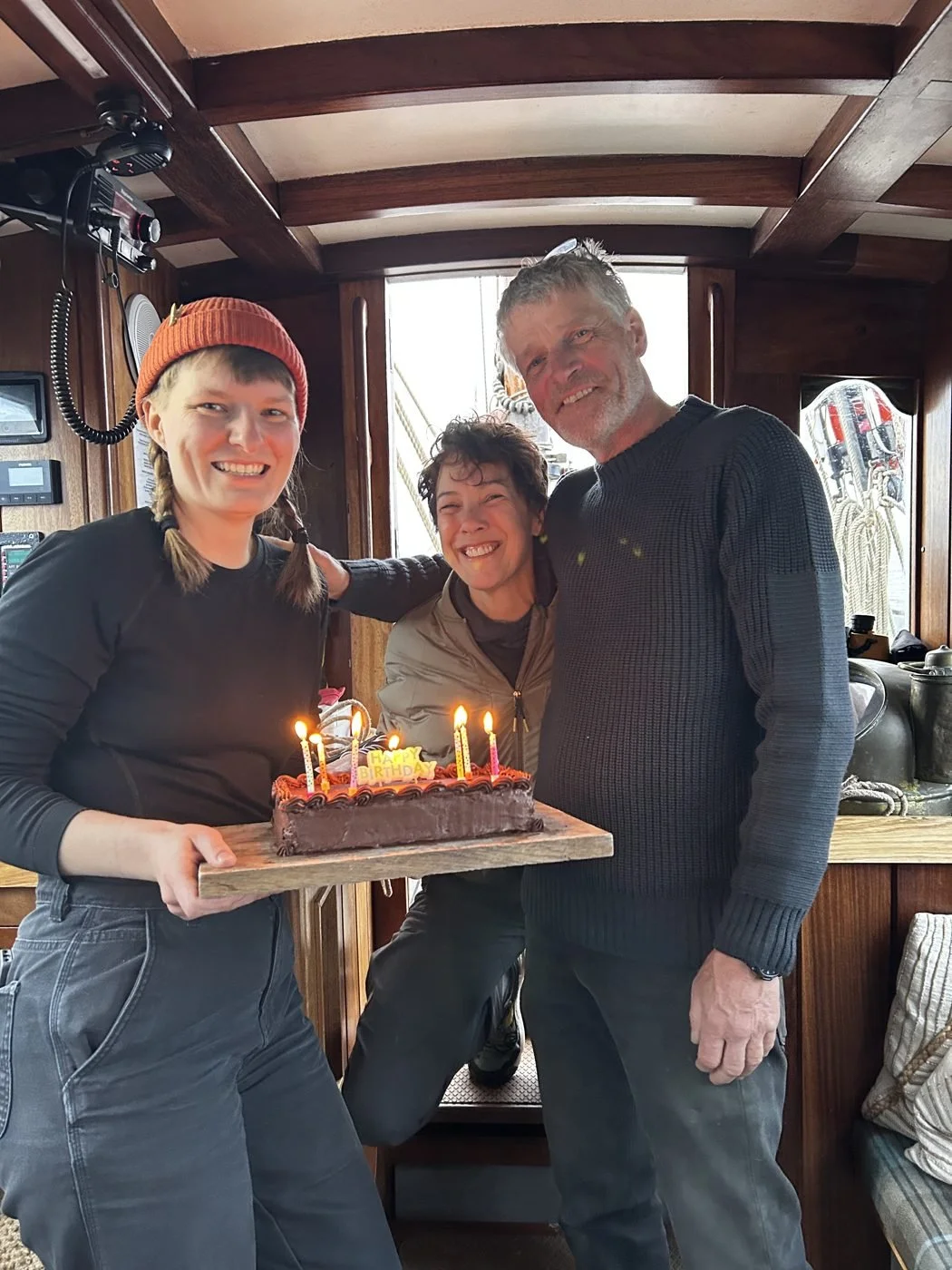 A young woman is holding a birthday cake with lit candles, with two older people, a woman and a man, standing beside her in a cozy wooden interior, celebrating a birthday.