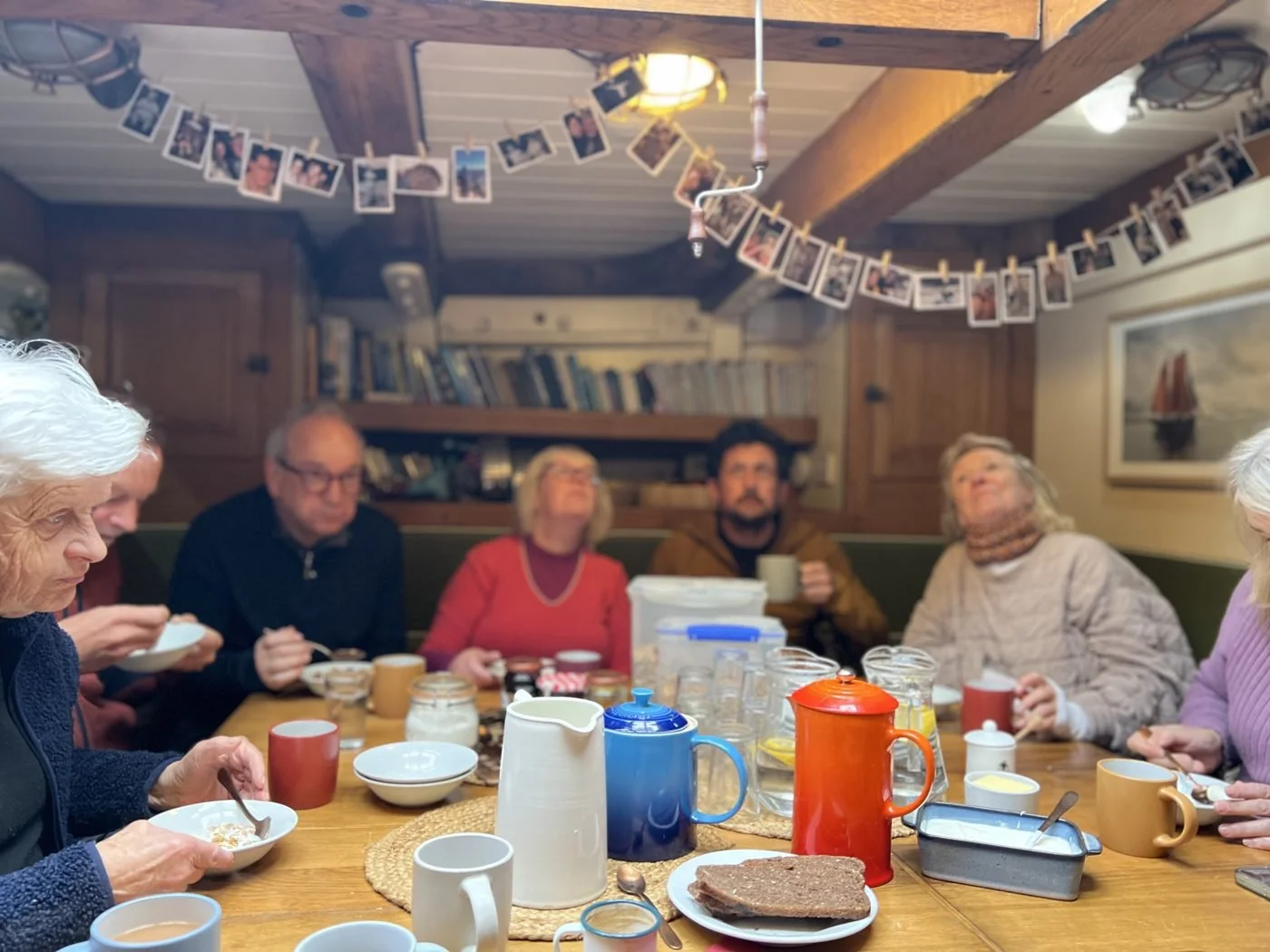 A group of elderly people sitting around a dining table enjoying a meal in a cozy, wood-paneled room. The table has various mugs, bowls, and a plate with bread. Photos are hanging on a string from the ceiling, and a bookshelf is in the background.