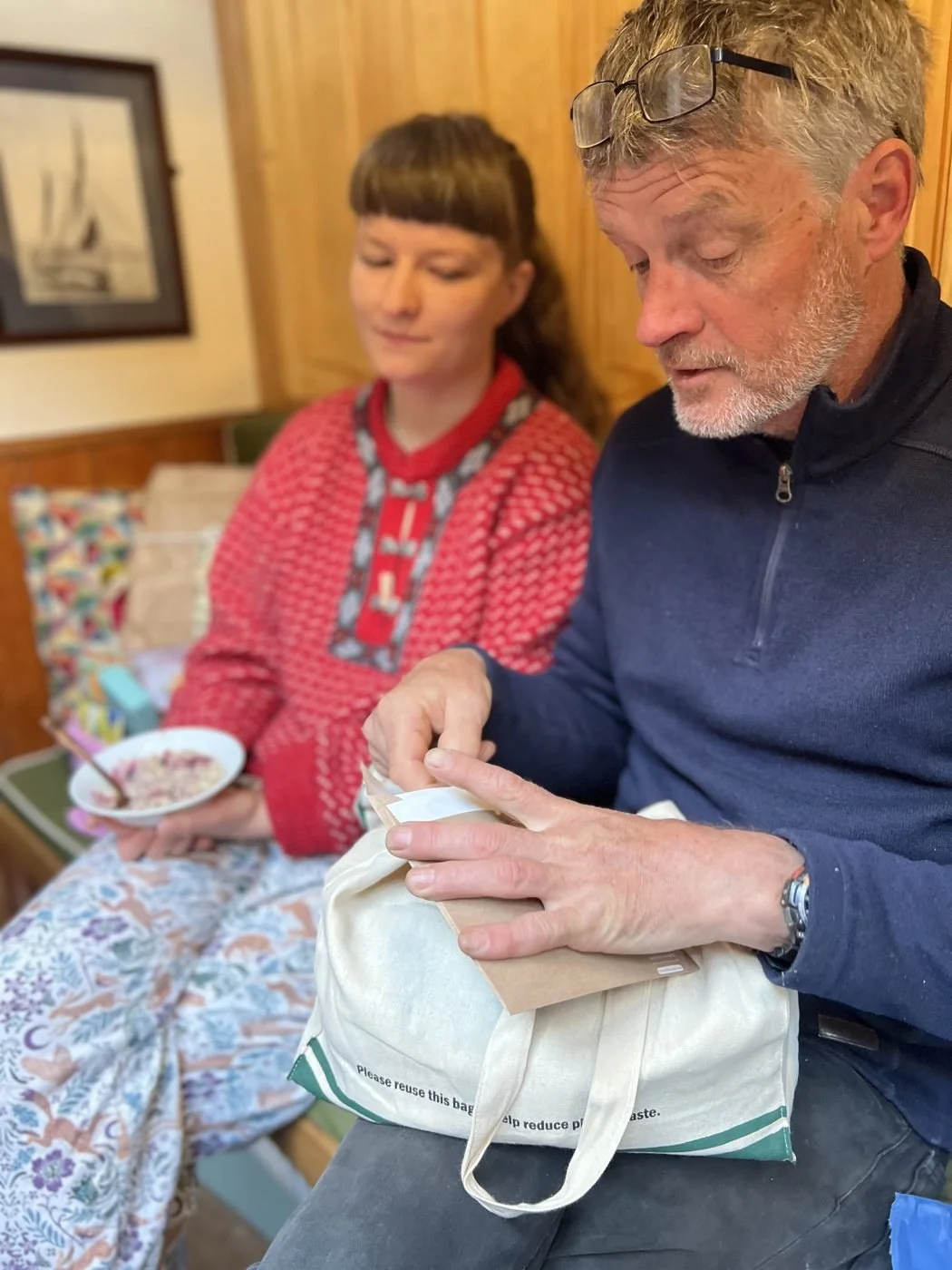 A man with glasses on his head is opening a paper bag, sitting next to a woman with a bowl of cereal, in a room with wooden walls.
