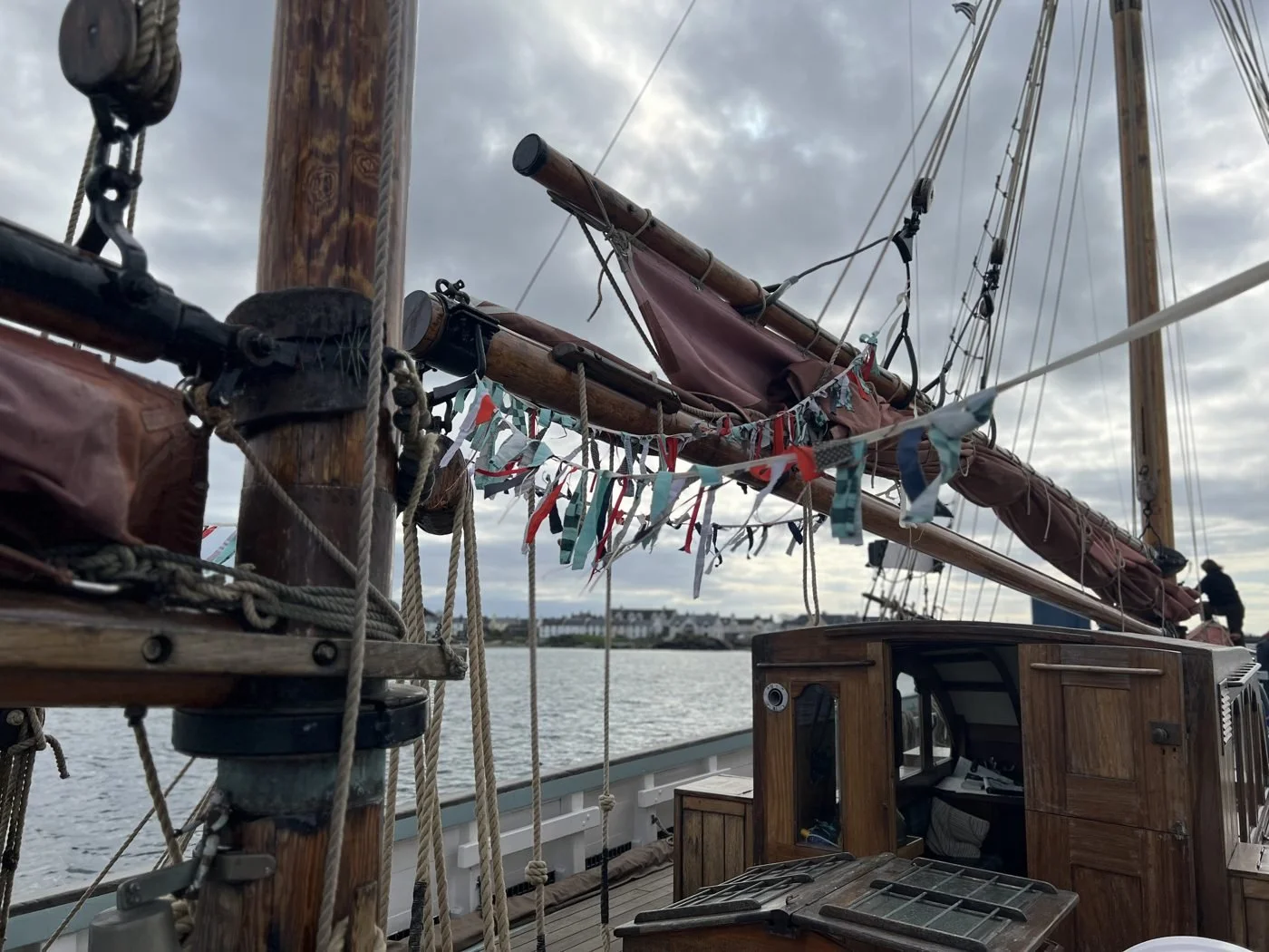 View of a sailing ship's wooden mast, rigging, and sails, with colorful ribbons hung along the ropes. A person is visible in the background near the ship, with water and shoreline under cloudy sky.