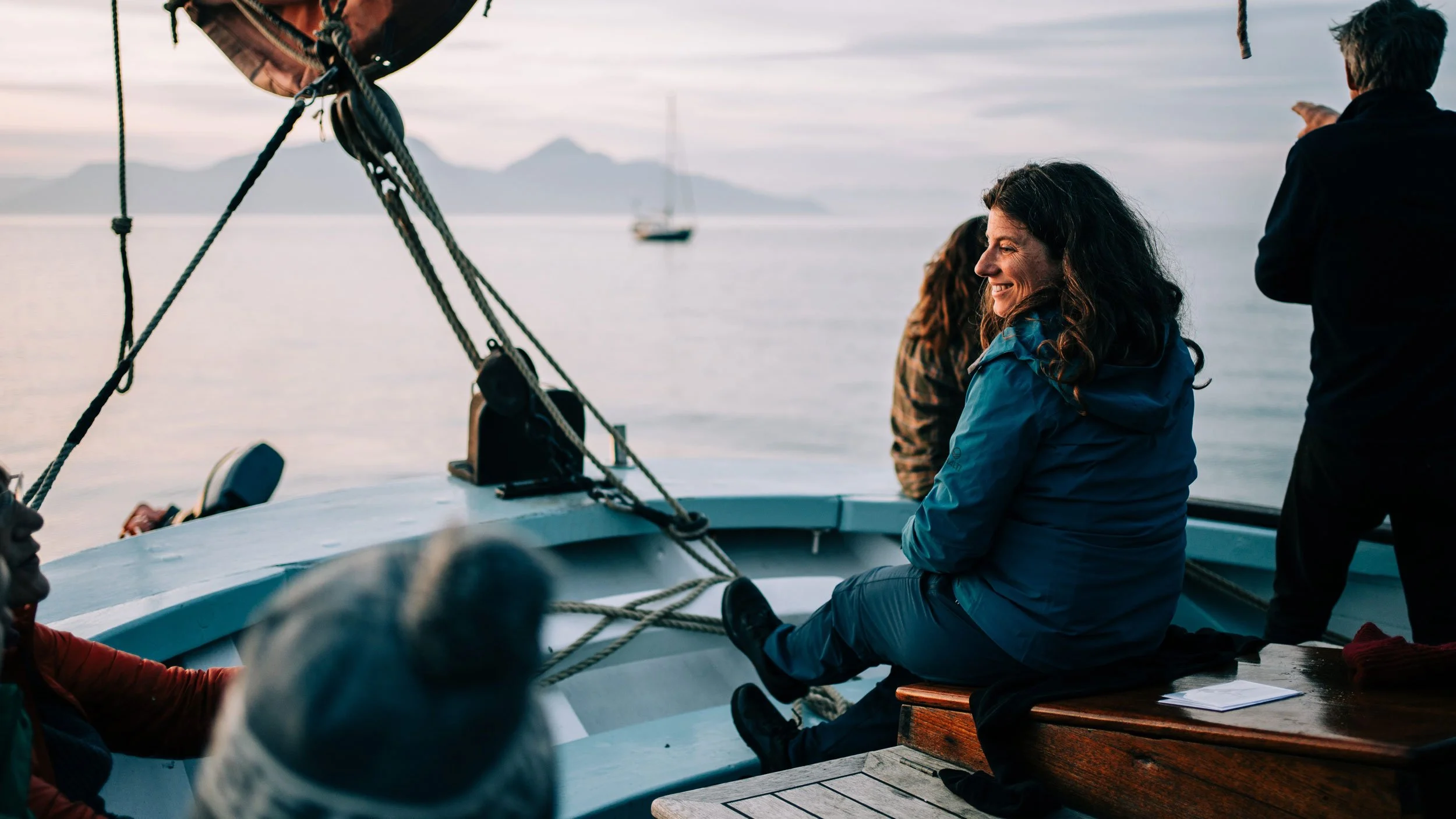People sitting on a boat deck with a woman smiling, wearing a blue jacket, with ocean and mountains in the background.