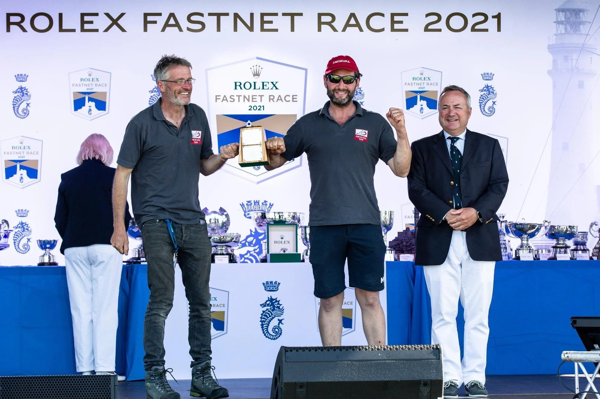 Three men standing on a stage during the Rolex Fastnet Race 2021 awards ceremony. The man in the middle, wearing sunglasses and a red cap, is celebrating with a fist pump and holding a trophy. The man on the left is holding the trophy's frame, smiling. The man on the right is dressed in a blazer and white pants, smiling. The background features a large white banner with the event name and trophies on a table behind them.