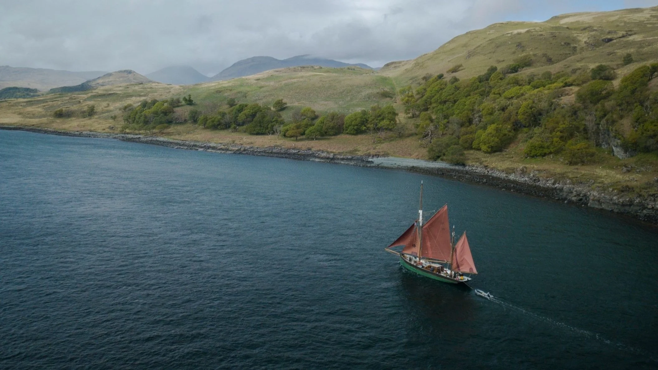 A sailing ship with red sails on a body of water near a green, hilly shoreline with mountains in the background.