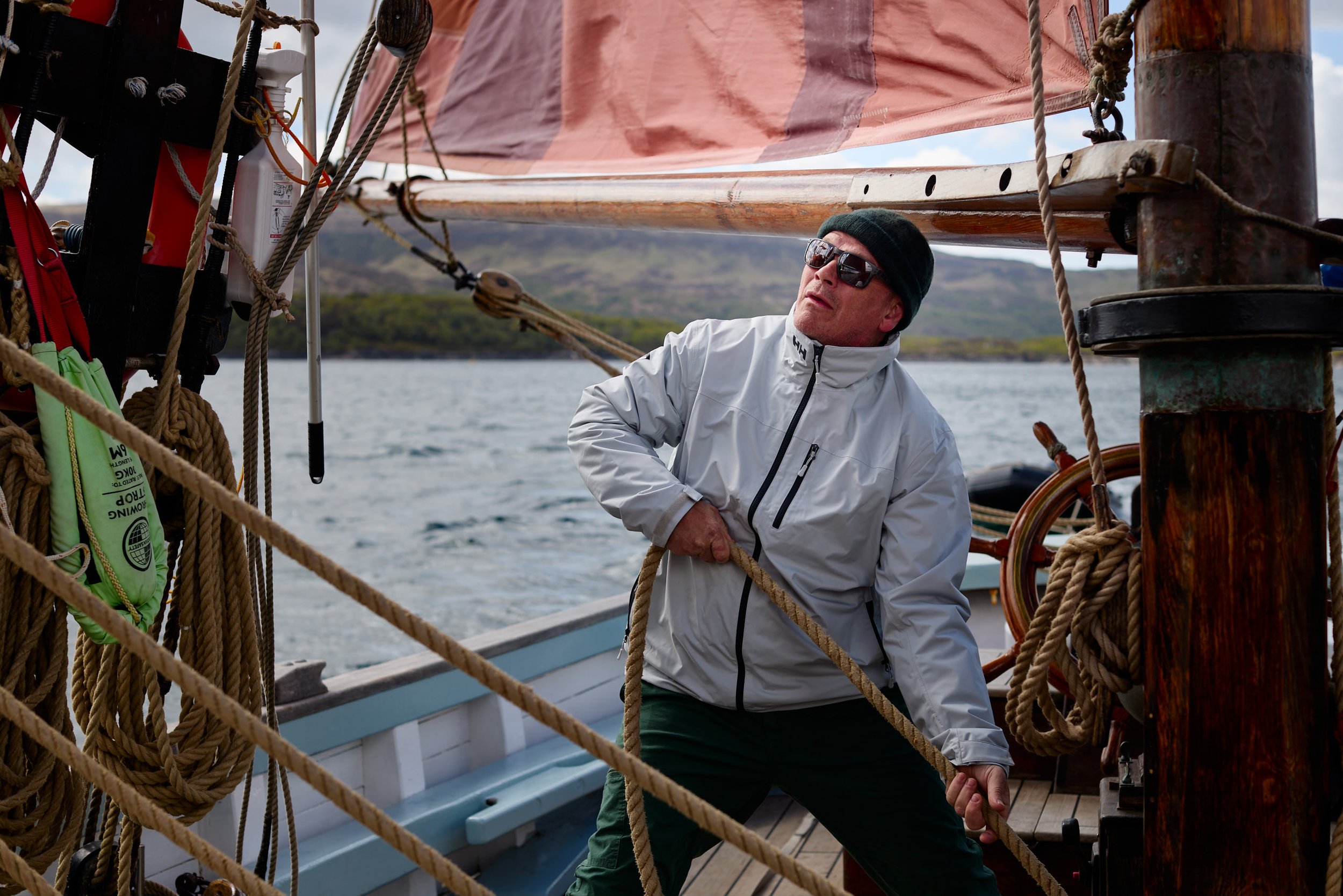 A person wearing sunglasses, a black beanie, and a white jacket standing on a sailboat, surrounded by ropes and equipment, with a scenic water and land background.