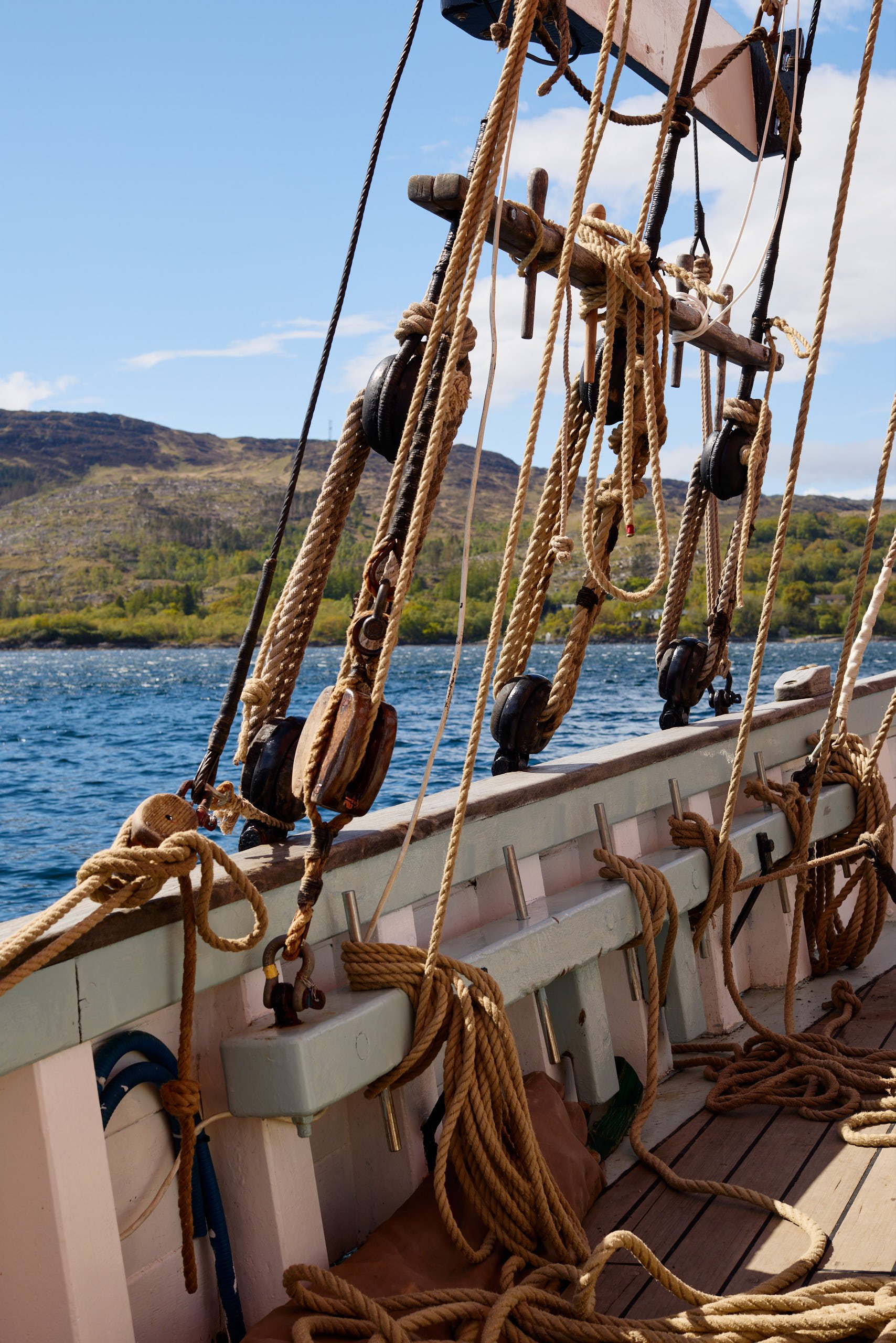 Ropes and pulleys on the deck of a sailing ship with a scenic landscape of hills, water, and blue sky with clouds in the background.