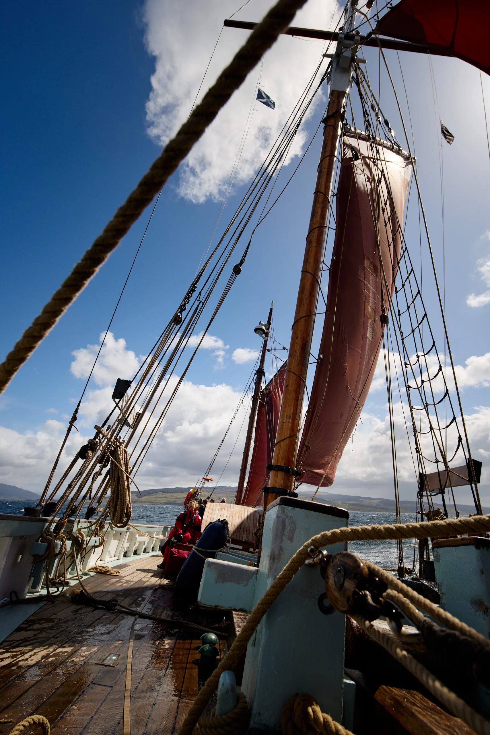 View from the deck of Provident  with a large red sail, wooden mast, and rigging ropes, with a blue sky and clouds in the background. Some people are seated on the deck near the stern.