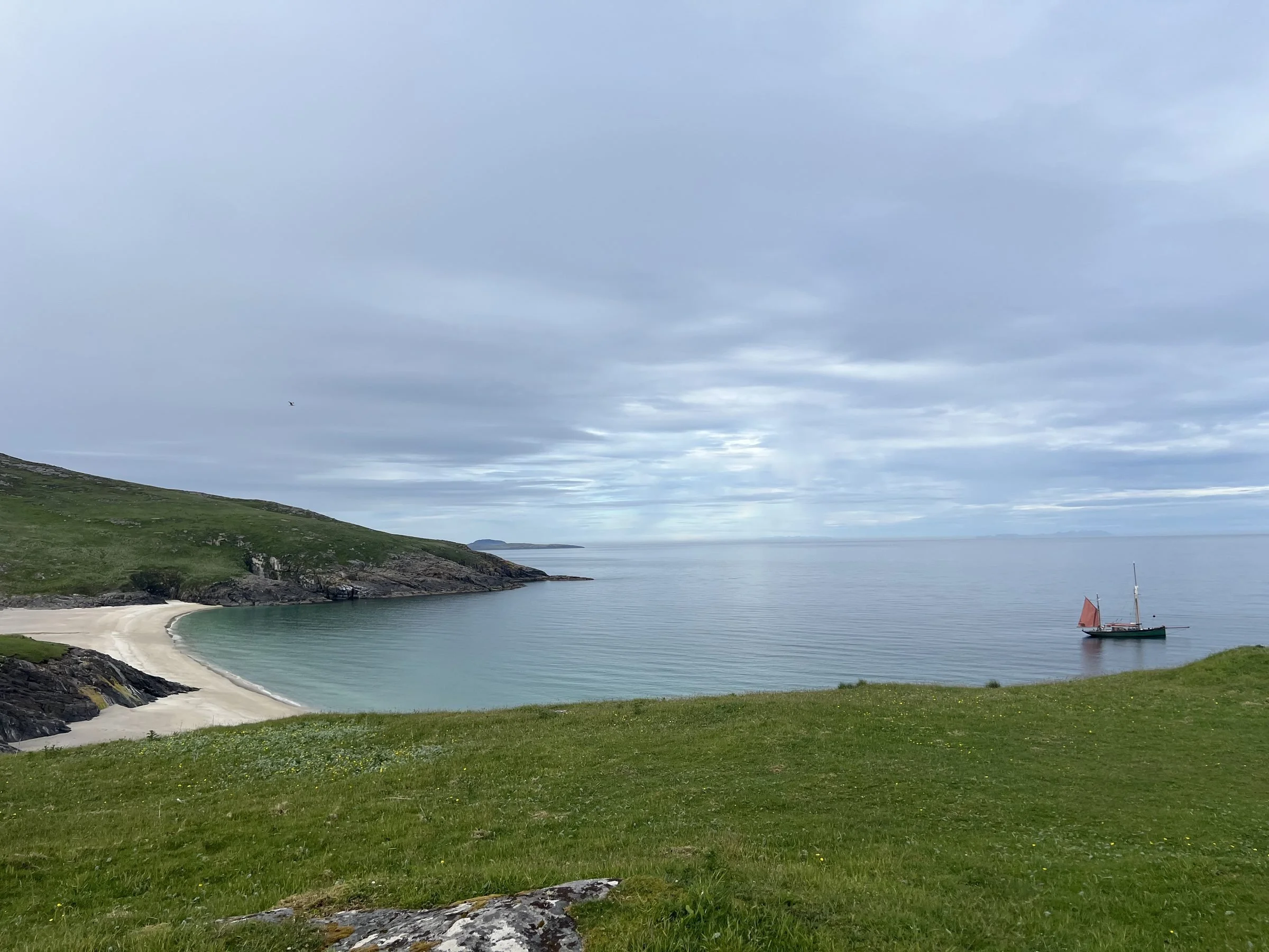 Scenic view of a coastal landscape with grassy hills, a sandy beach, calm water, and Provident with red sails under cloudy sky on the Island of Barra