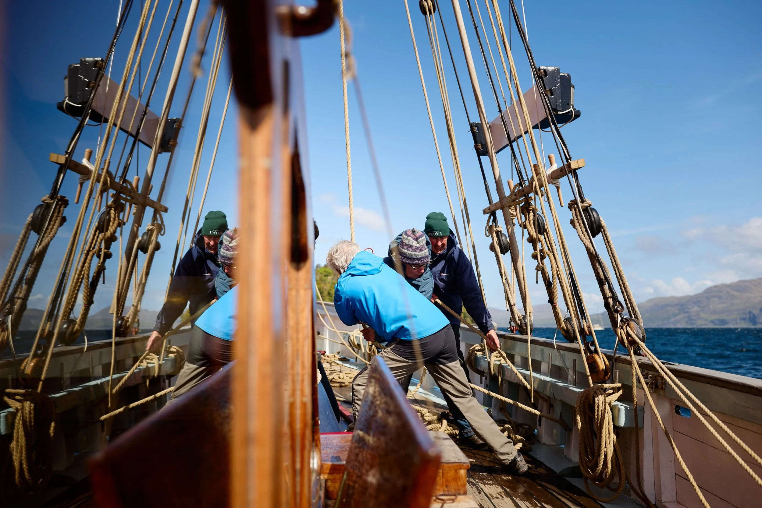 People working together on the deck of a wooden sailing ship with a clear blue sky and mountains in the background.