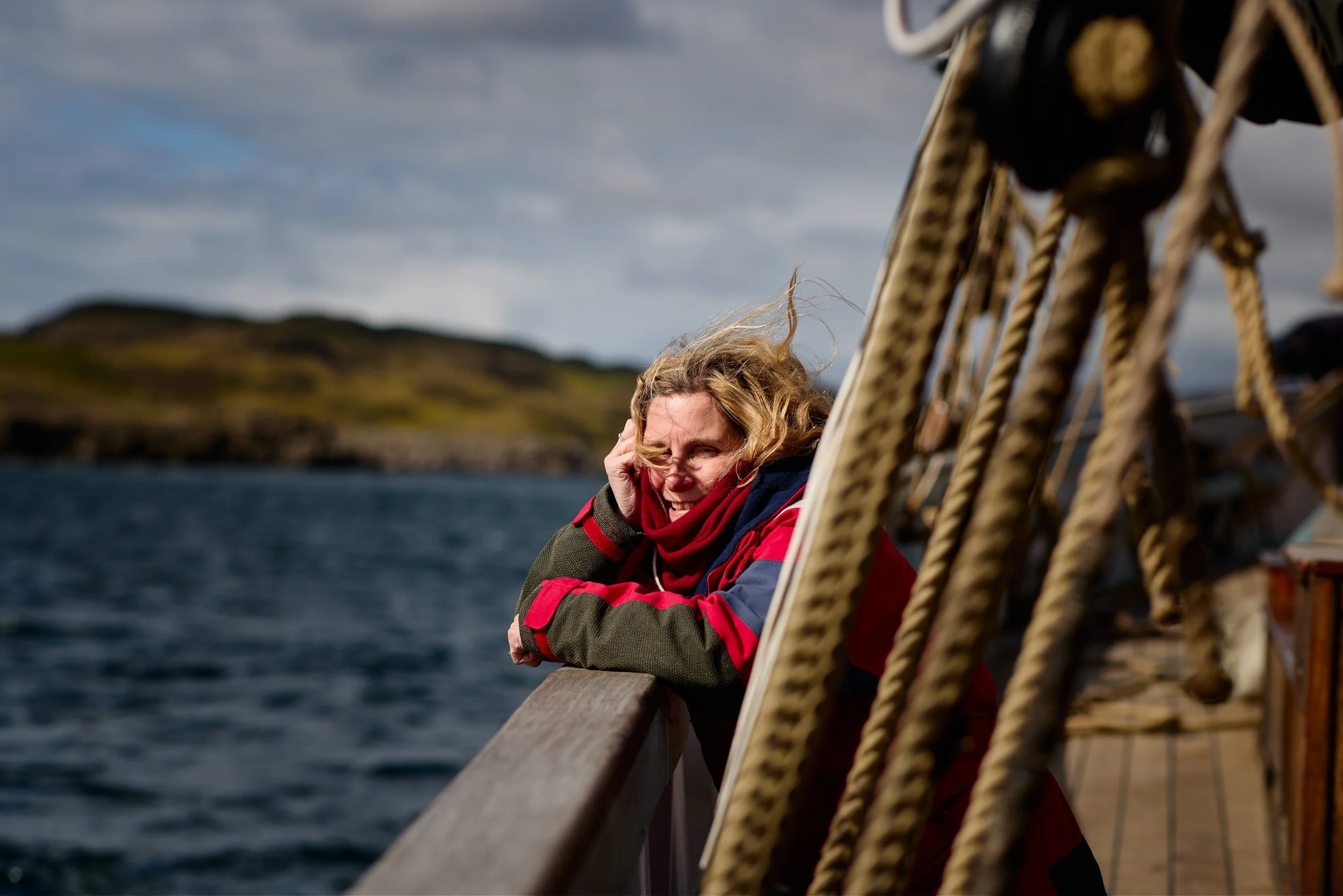 A woman with curly blonde hair and a red and gray jacket looking pensive on a boat, with ropes and the ocean in the background and a cloudy sky.