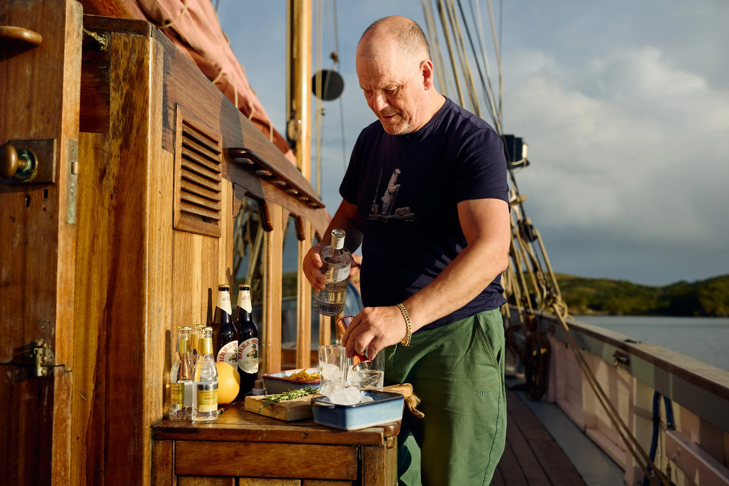 A man with a shaved head and a beard, wearing a navy t-shirt and green shorts, is preparing drinks at an outdoor bar on a boat or ship. The bar is made of wood and has bottles, lemons, and glasses on it. The man is holding a bottle and pouring a drink, with a cloudy sky in the background.