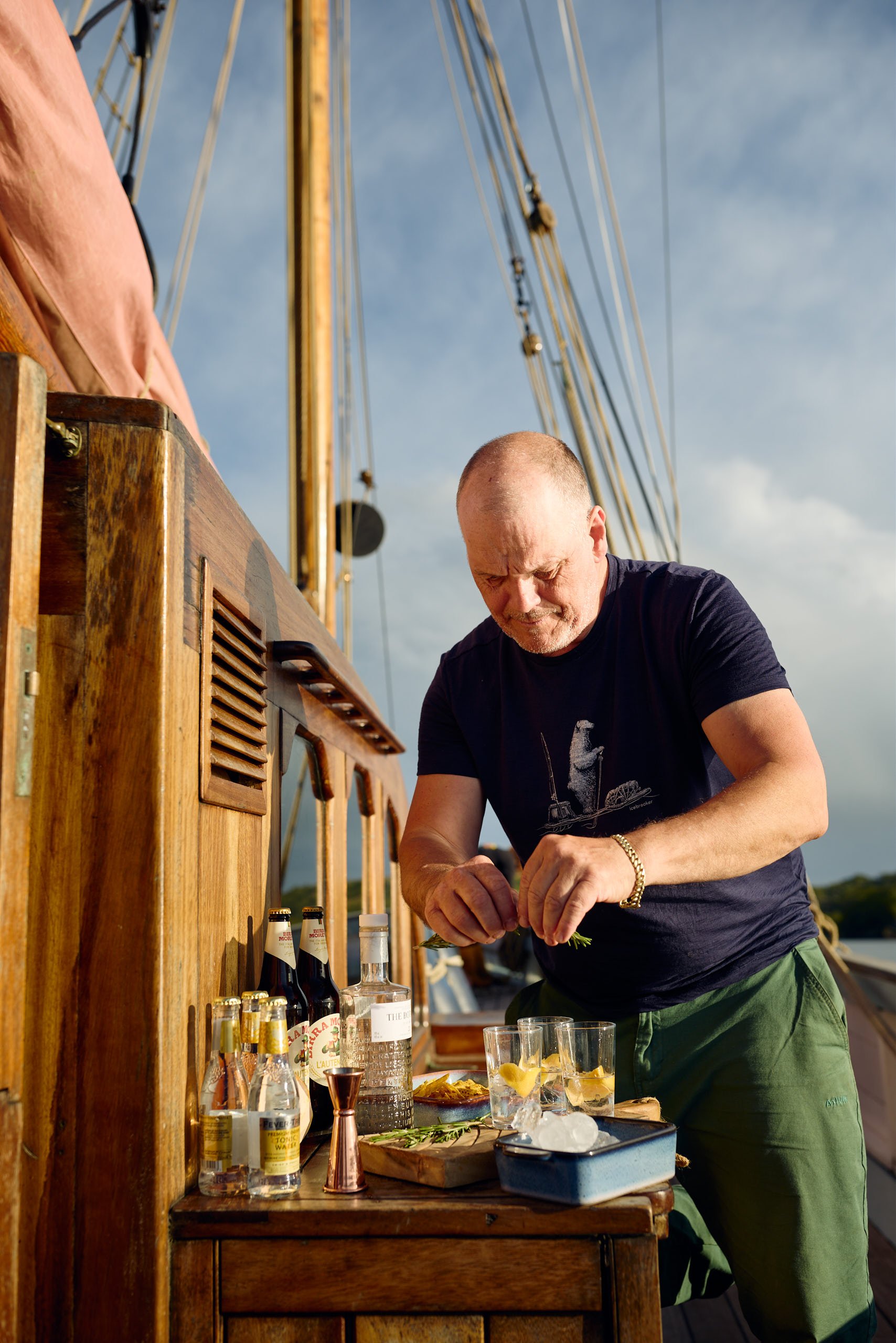 A man with a shaved head wearing a black t-shirt and green shorts preparing drinks on a wooden outdoor bar with bottles of alcohol and mixers, under a clear sky.
